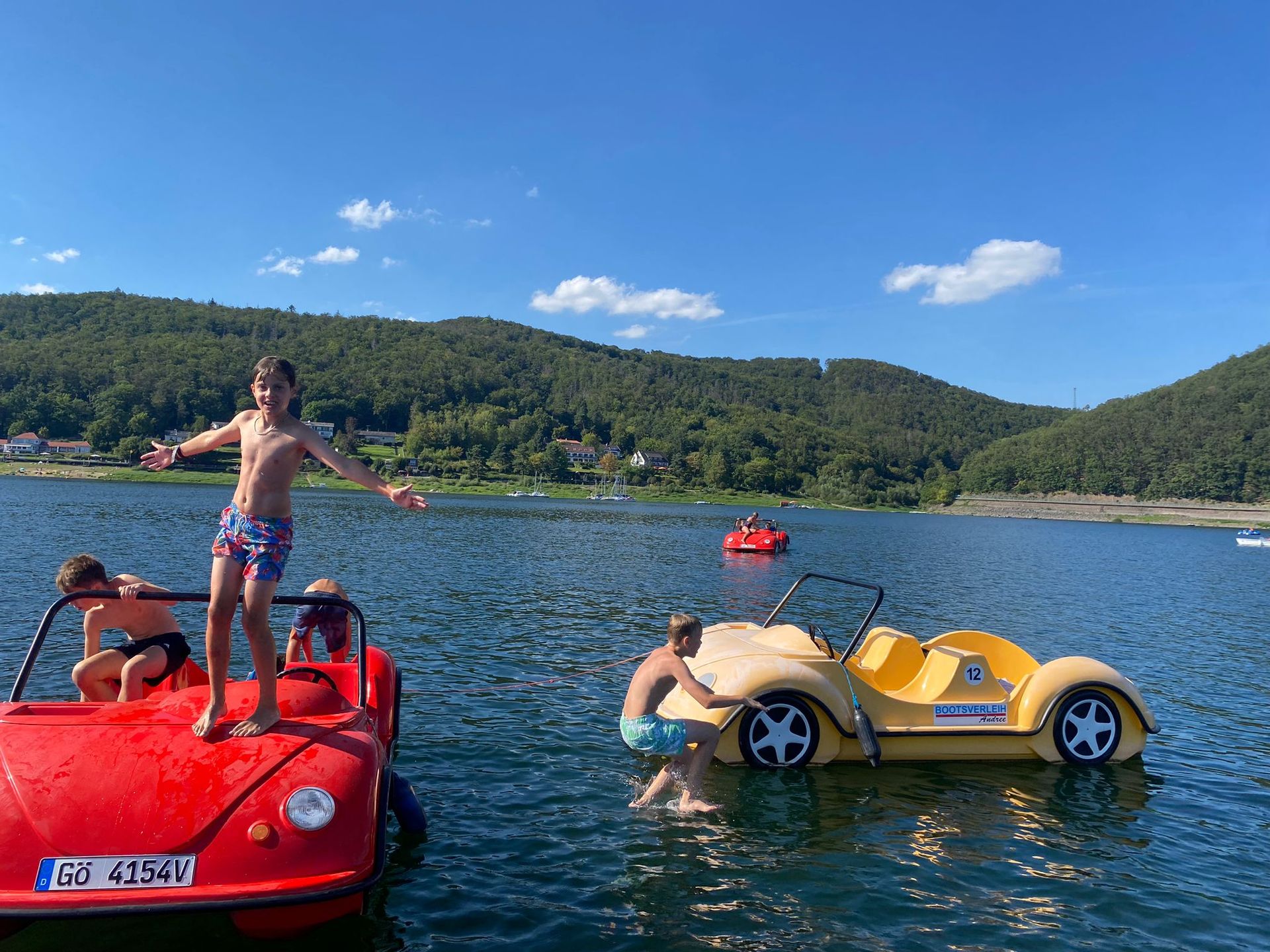 Drei Kinder auf Tretbooten in Form von Autos auf einem See, eines steht mit ausgestreckten Armen vor einem bewaldeten Hügel.