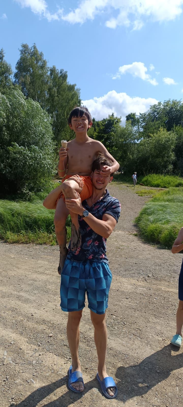 Eine Person trägt eine andere auf den Schultern im Freien. Sie befinden sich in der Nähe von Bäumen und der Himmel ist blau.