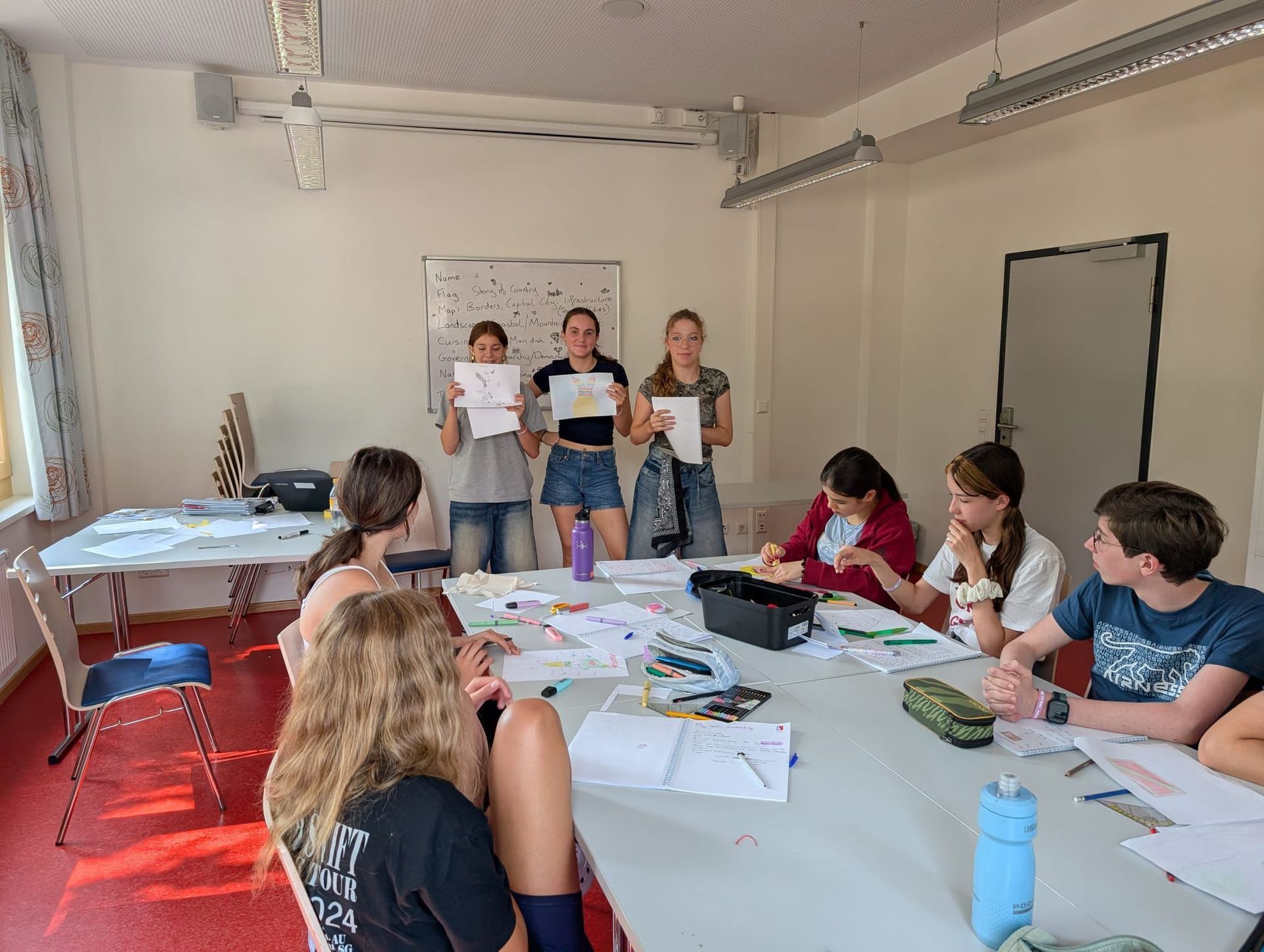 Schülerinnen in einem Klassenzimmer. Drei Mädchen präsentieren Zeichnungen, andere sitzen an einem Tisch und arbeiten an Projekten.