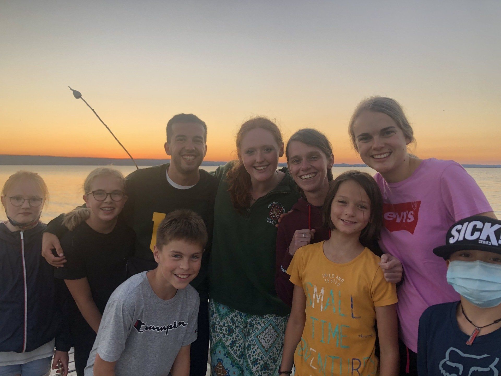 Eine Gruppe lächelnder Menschen sitzt bei Sonnenuntergang auf einem Pier; das Wasser ist ruhig, einige tragen Masken.