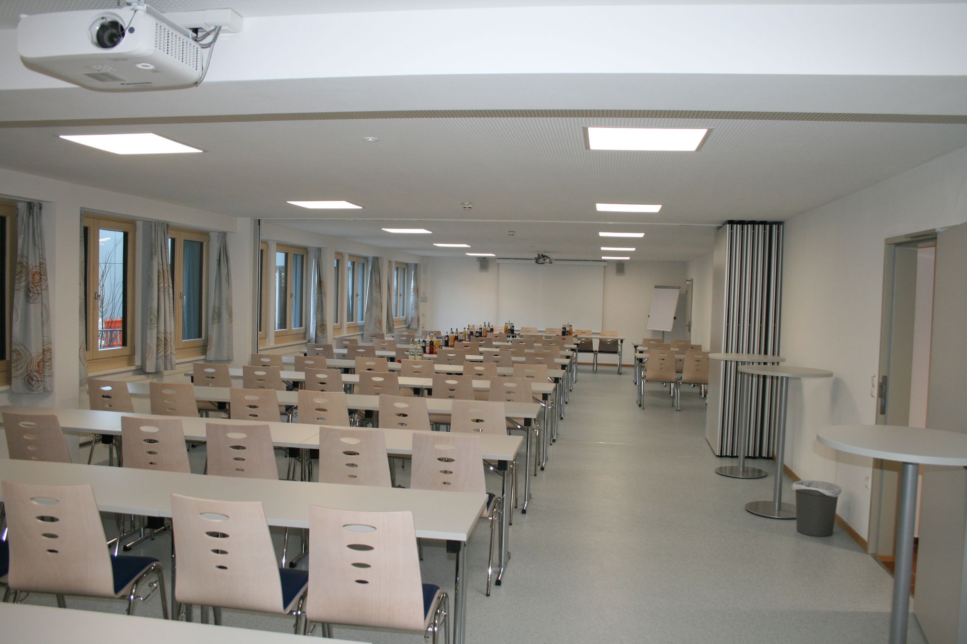 Klassenzimmer mit Reihen von Tischen und Stühlen, Projektor an der Decke.