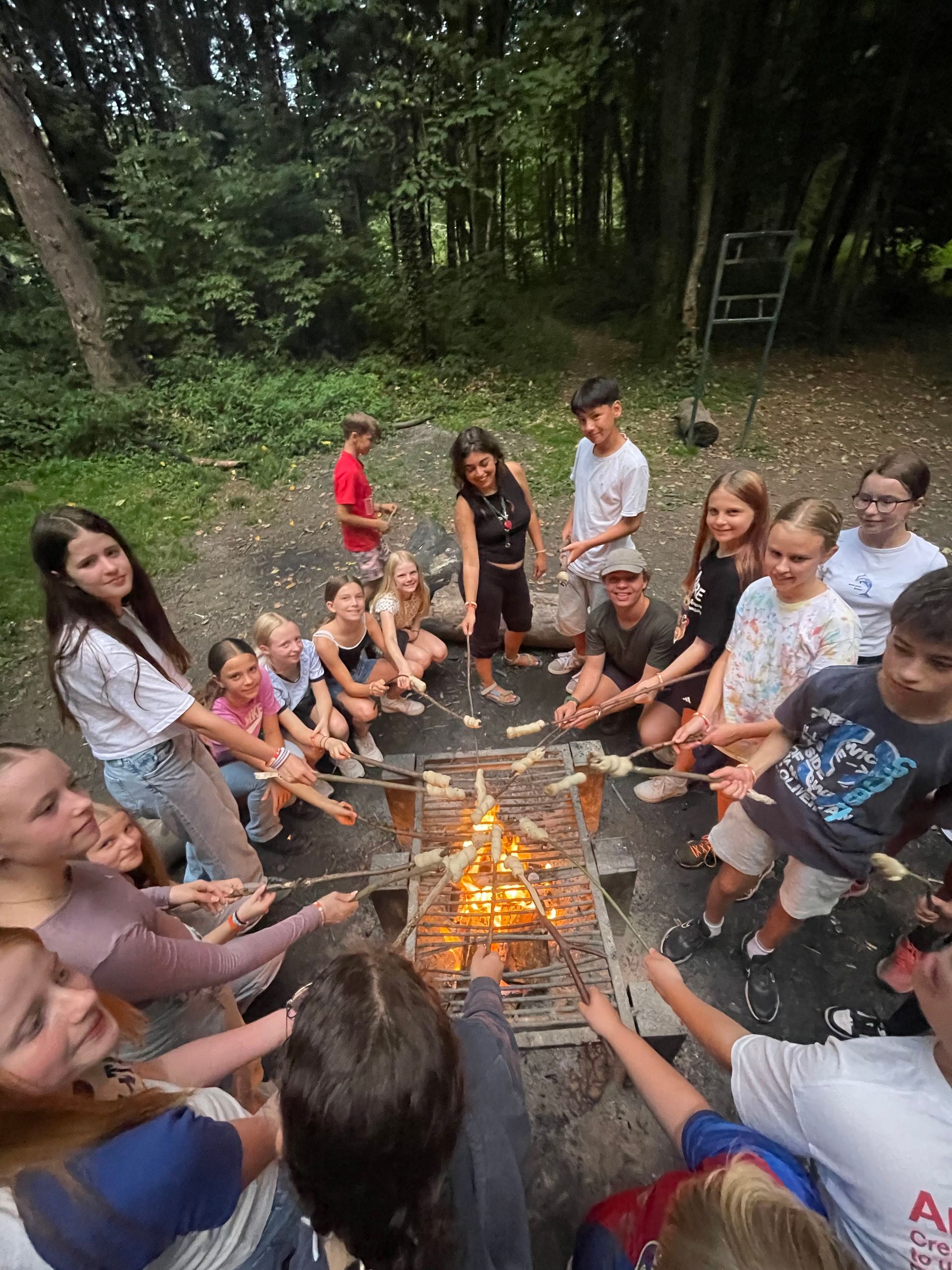 Eine Gruppe Kinder röstet Essen über einem Lagerfeuer in einem Waldgebiet.