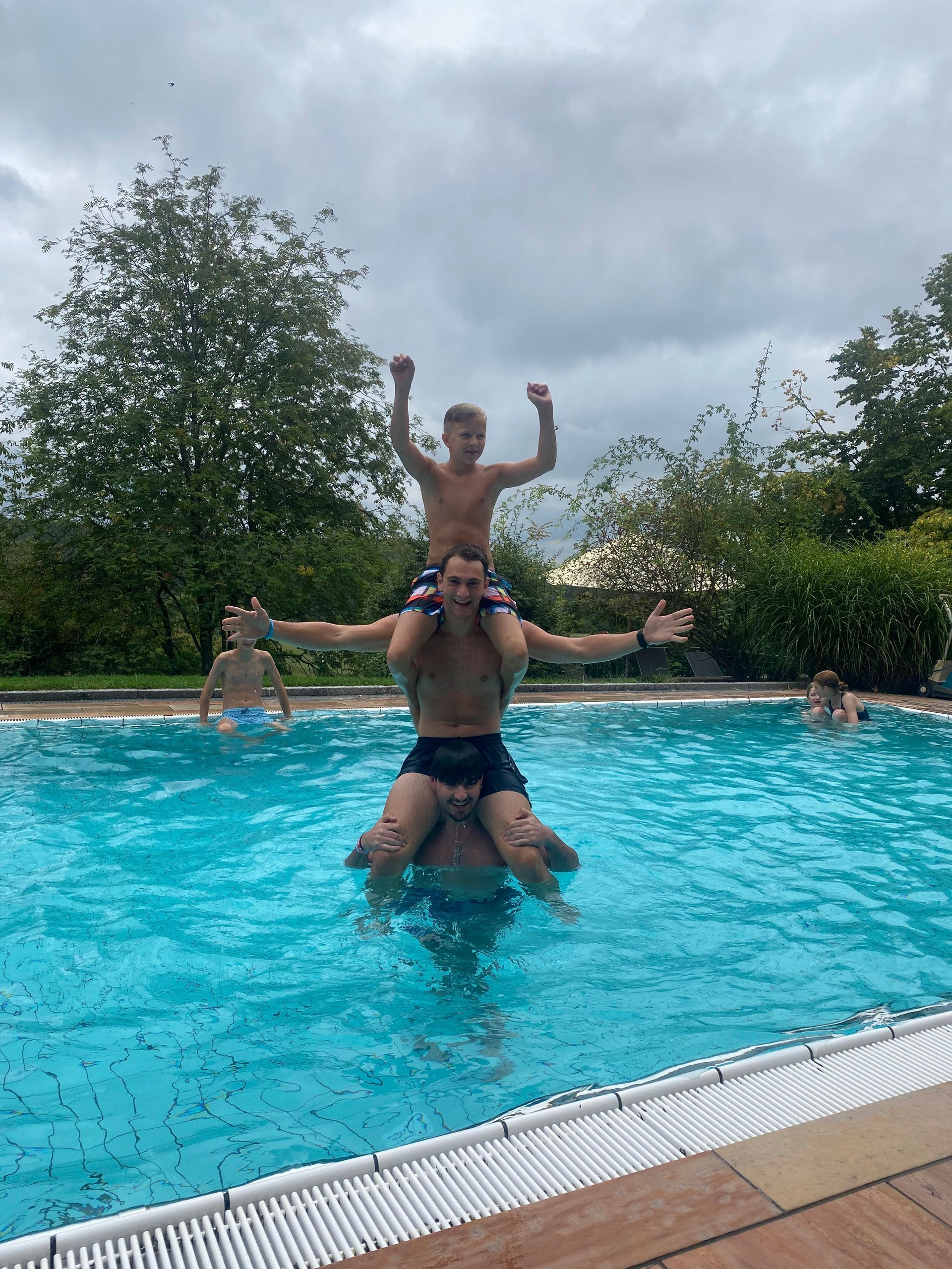 Menschen in einem Pool, einer auf den Schultern des anderen. Ein Kind oben hebt die Arme, bewölkter Himmel.
