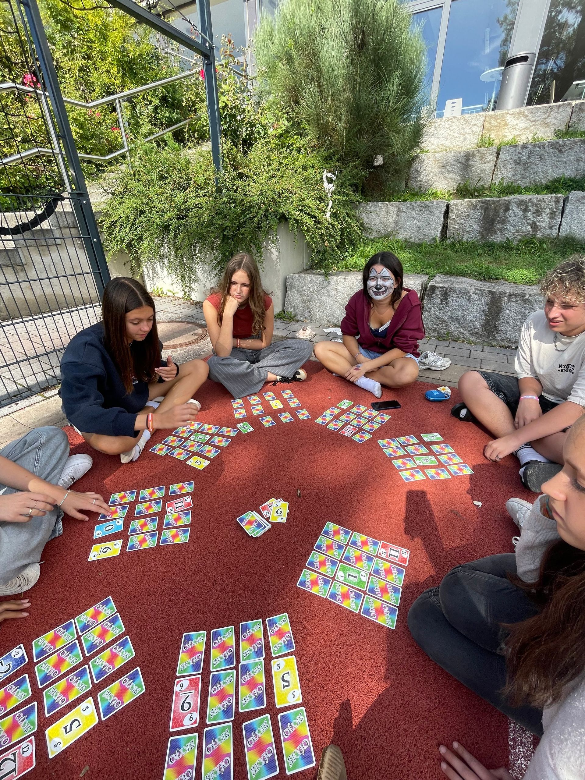 Eine Gruppe Teenager spielt an einem sonnigen Tag draußen auf einem roten Untergrund Karten.