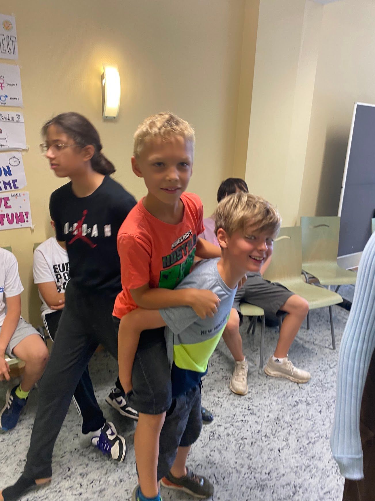 Zwei Jungen spielen Huckepack in einem Zimmer. Andere schauen zu, einer trägt eine Brille. Helles Licht.