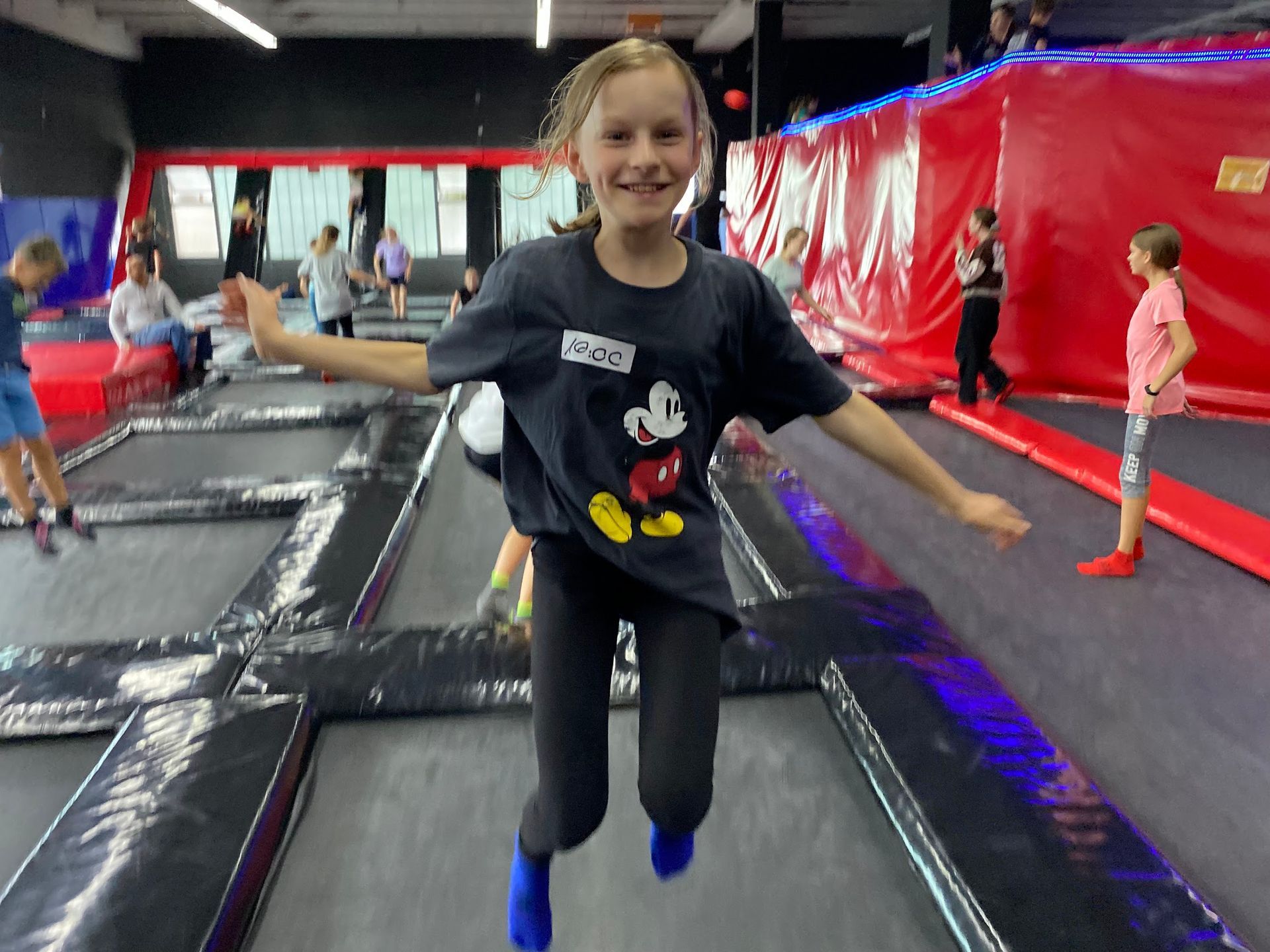 Mädchen in einem Mickey-Mouse-Shirt springt lächelnd mit ausgestreckten Armen auf einem Trampolin. Schwarzer und roter Hintergrund.