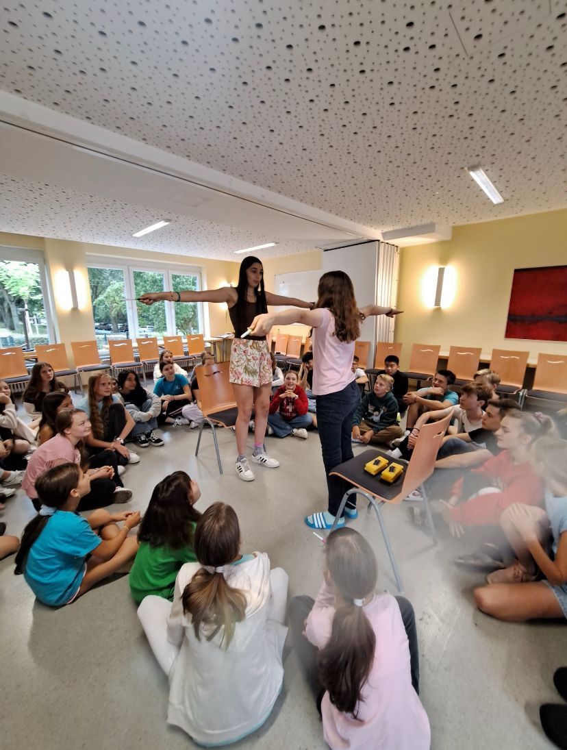 Zwei Mädchen demonstrieren einer Gruppe von Kindern, die in einem hell erleuchteten Raum im Kreis sitzen, ein wissenschaftliches Experiment.