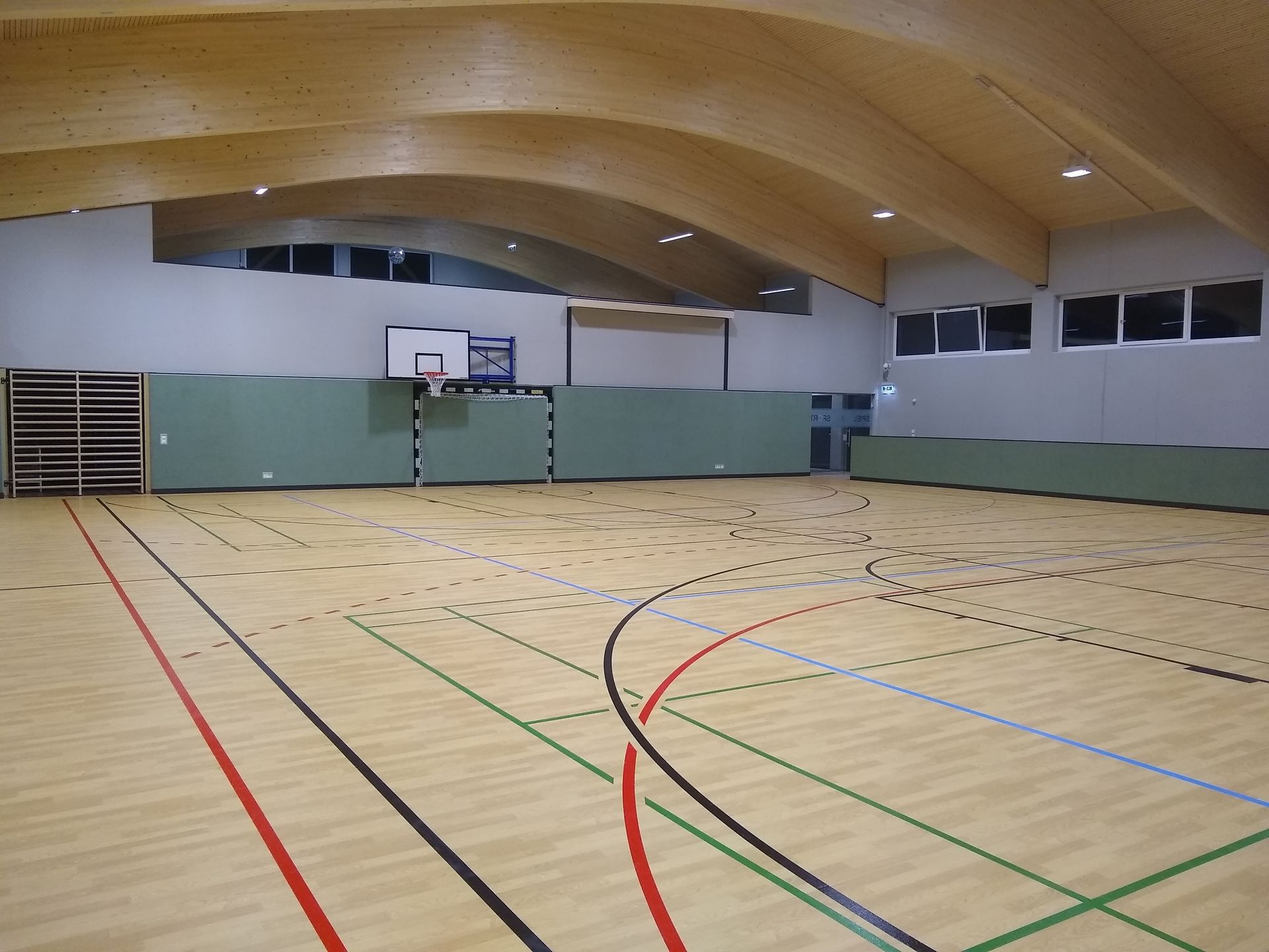 Turnhalle mit Holzboden und -decke, Basketballkorb und aufgemalten Spielfeldlinien.