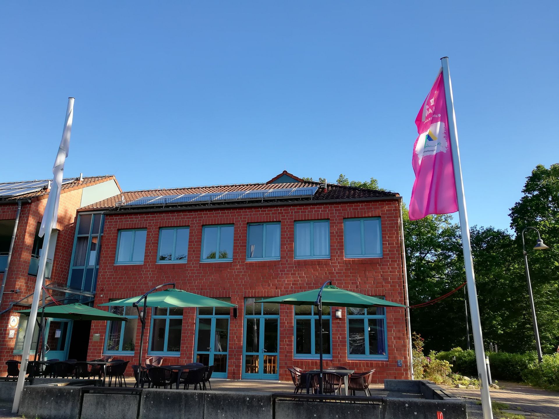 Backsteingebäude mit Solarpaneelen, türkisfarbenen Türen und Fensterrahmen, Sonnenschirmen und Fahnen vor blauem Himmel.