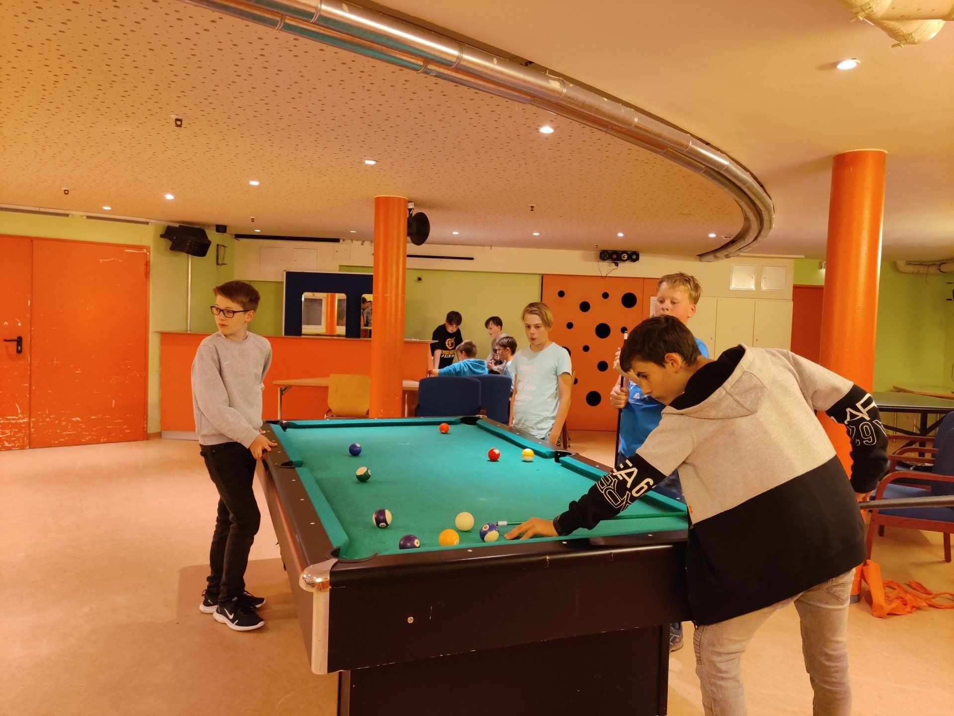 In einem orange-grünen Raum spielen Leute Billard. Einer will gerade den Stoß ausführen, die anderen schauen zu.