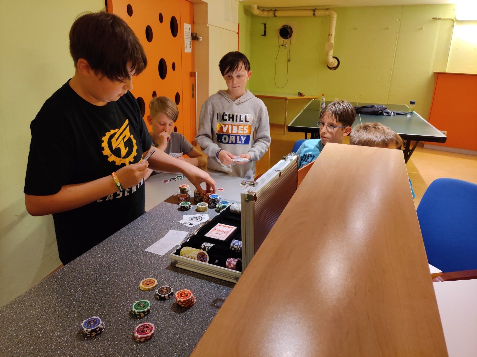 Kinder spielen in einem Raum mit orangefarbenen und grünen Wänden ein Kartenspiel mit Pokerchips auf einem Tisch.