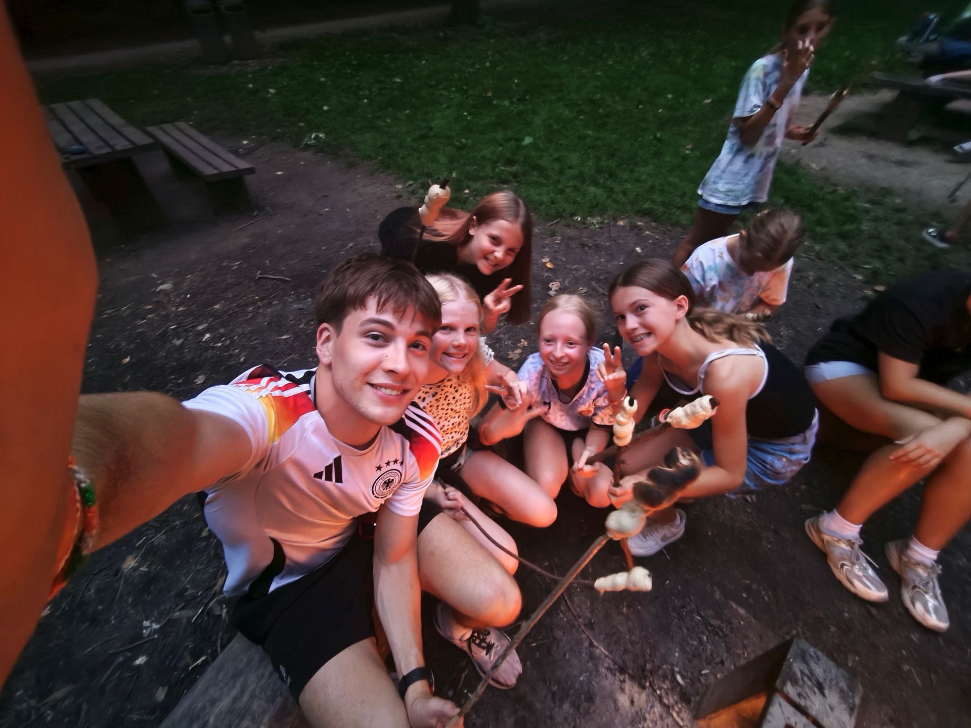 Eine Gruppe von Menschen sitzt um ein Lagerfeuer und grillt Essen. Sie lächeln und zeigen Friedenszeichen. Draußen in der Abenddämmerung.