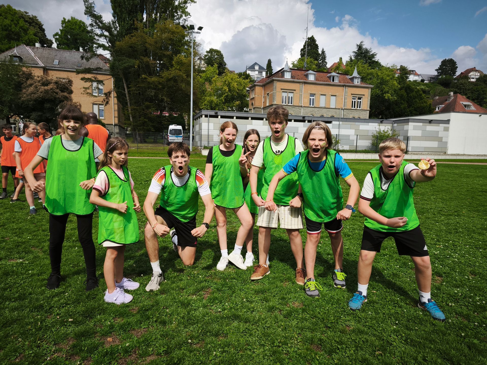Kinder in grünen Westen auf einer Wiese, die aufgeregte Gesichter machen; im Hintergrund Gebäude und Bäume.
