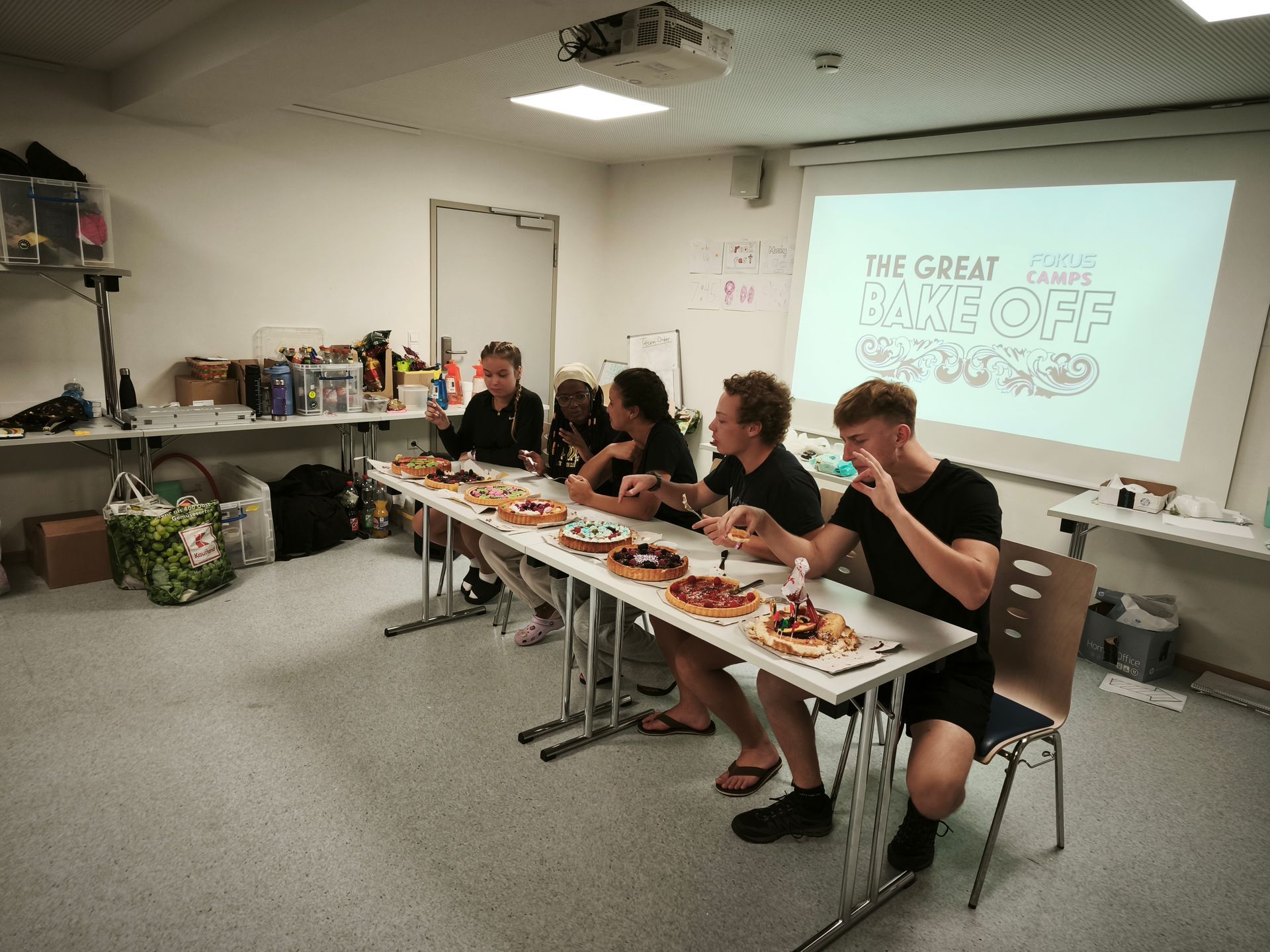 Menschen essen Gebäck an einem Tisch. „Das große Backen“ wird auf eine Leinwand projiziert.