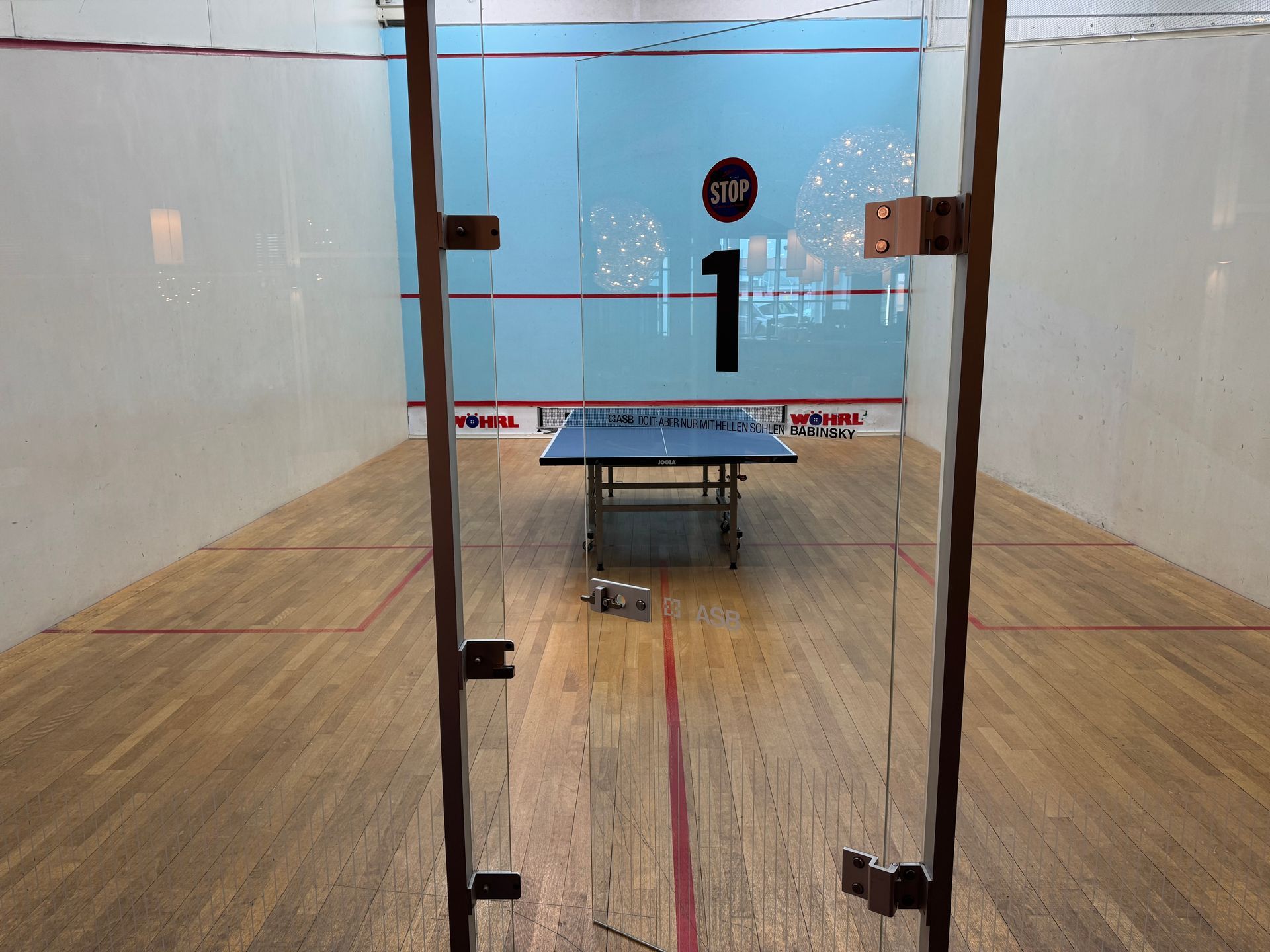 Squashcourt mit integrierter Tischtennisplatte. Holzboden mit Glastüren. Die Nummer 1 befindet sich an der Rückwand.