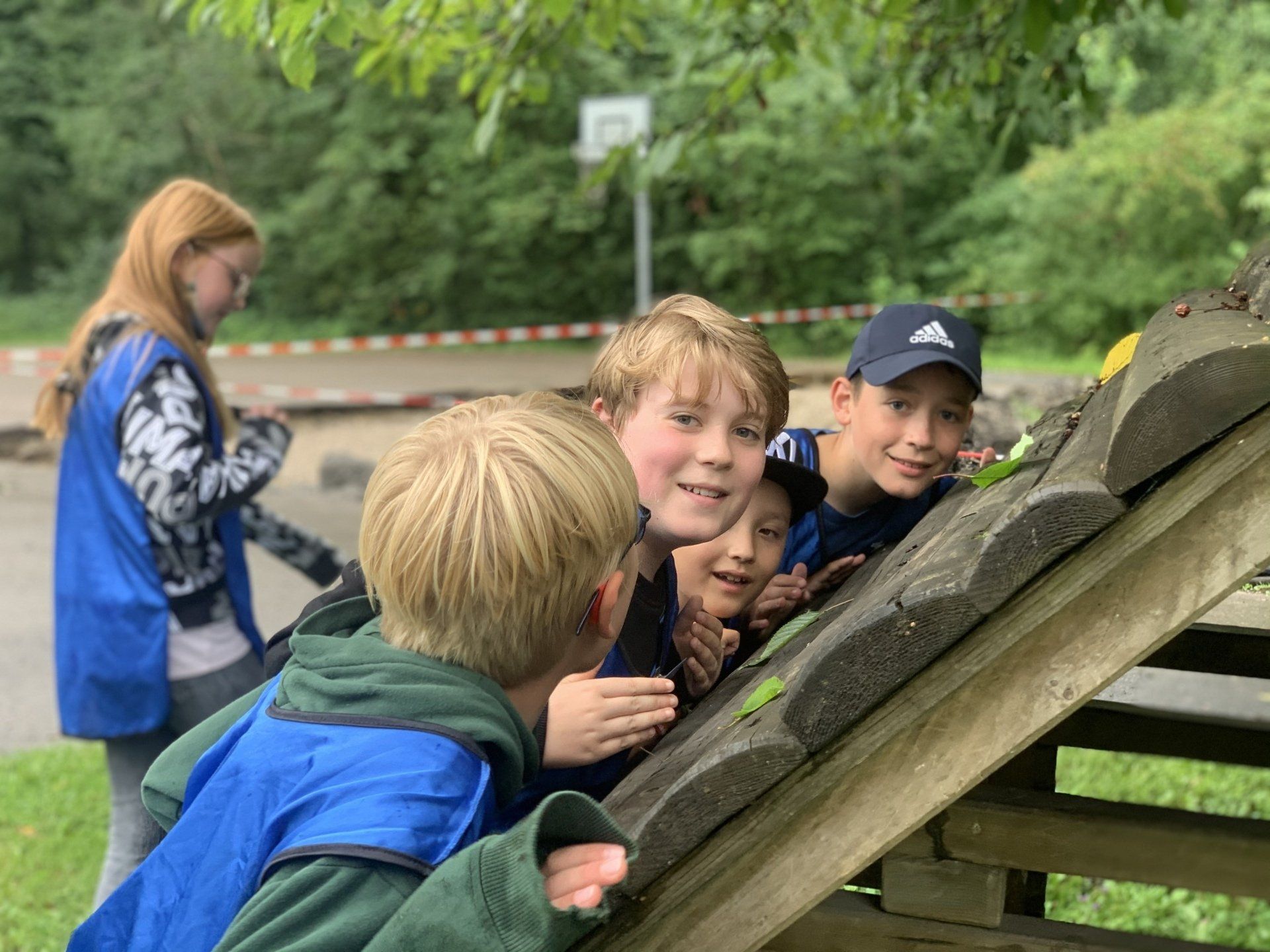 Eine Gruppe Kinder untersucht im Freien eine Holzkonstruktion. Ein Mädchen im Hintergrund trägt eine blaue Weste.