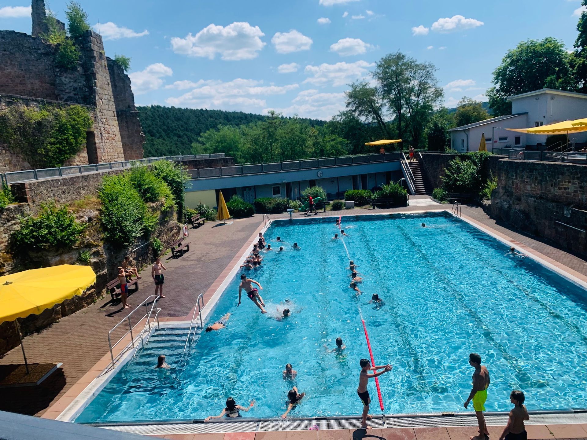 Freibad mit Menschen, neben einer Steinmauer unter blauem Himmel.