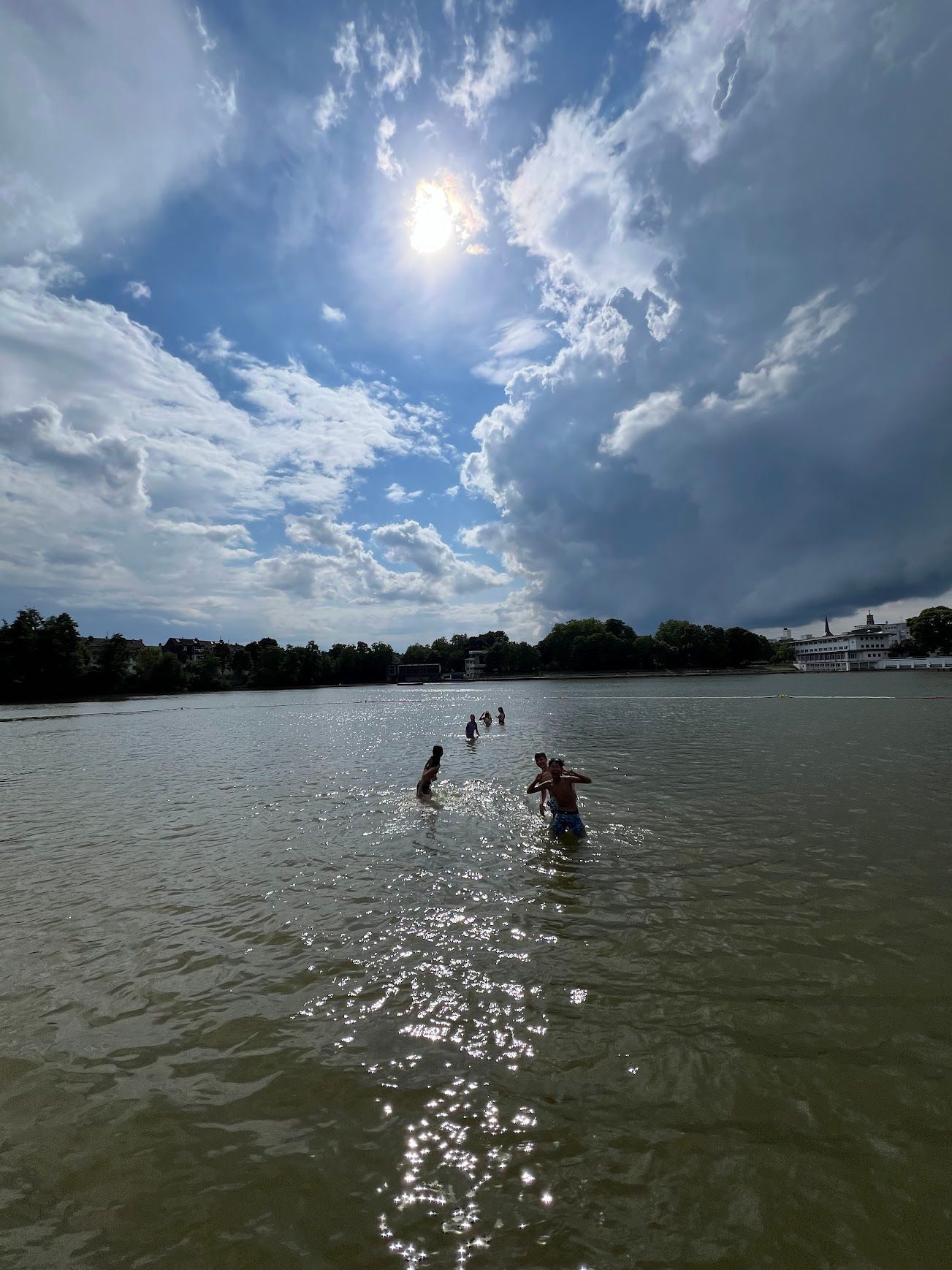 Menschen waten im Wasser in Richtung einer Baumgrenze bei strahlendem Sonnenschein und bewölktem Himmel.