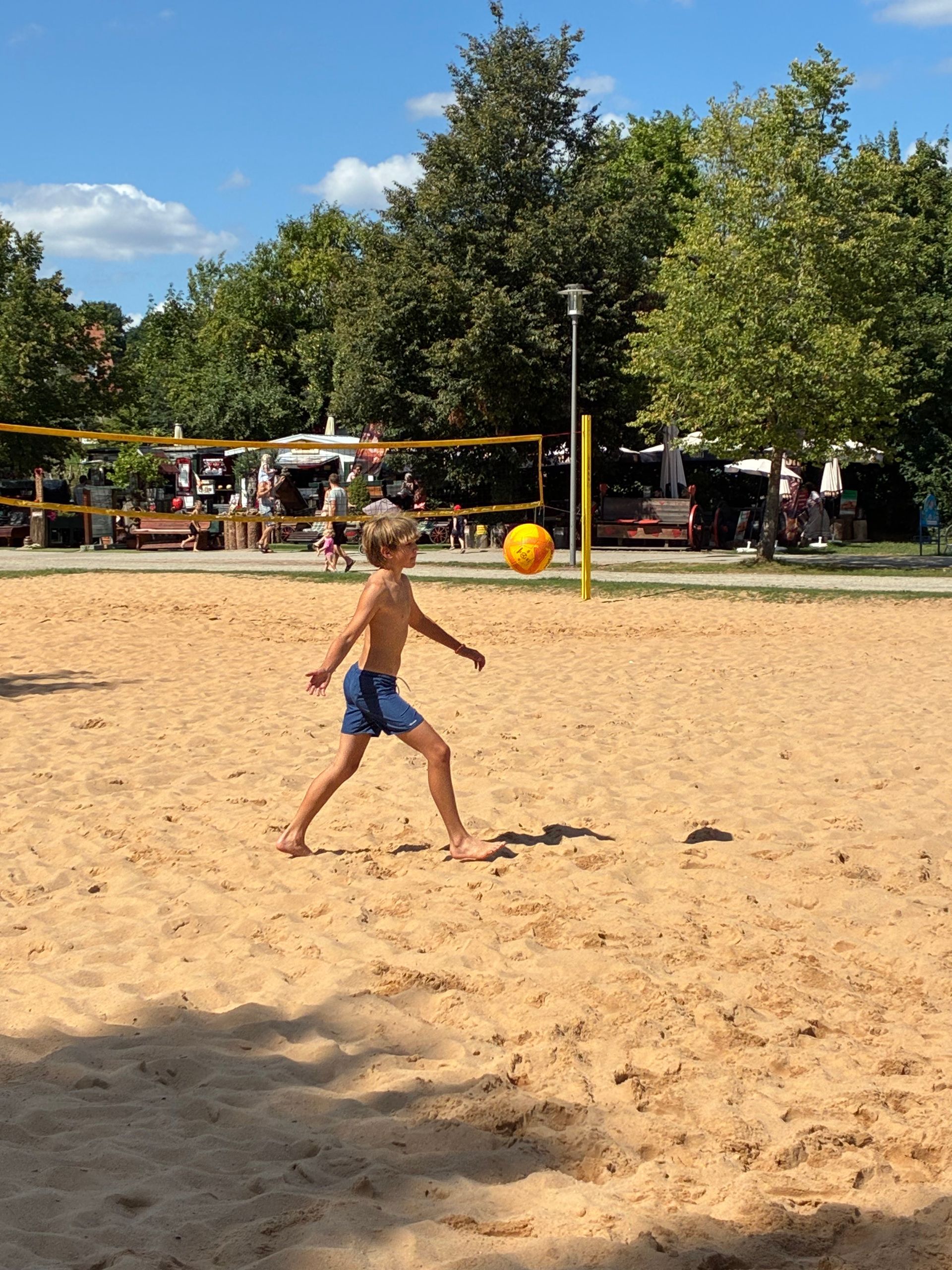 Ein Junge in blauen Shorts spielt Volleyball auf einem Sandplatz; der Ball befindet sich in der Luft. Im Hintergrund sind weitere Personen und Bäume zu sehen.