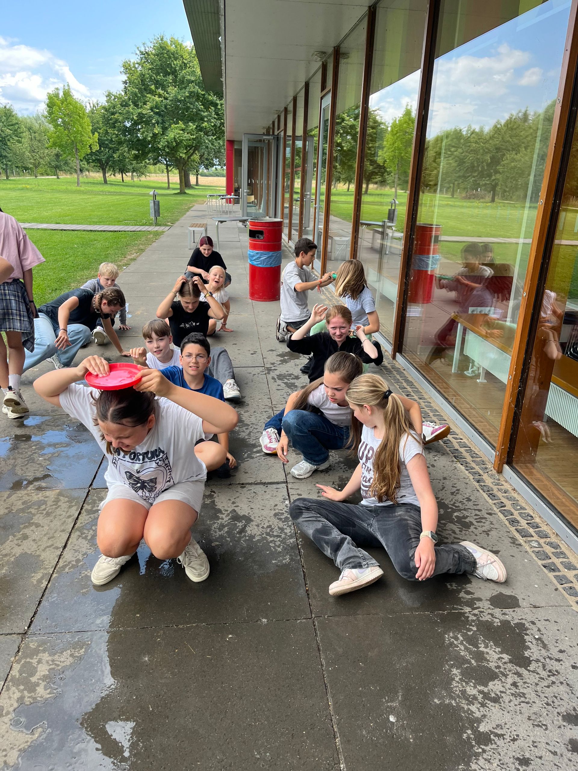 Kinder hocken und krabbeln draußen und balancieren Teller auf ihren Köpfen in der Nähe eines Gebäudes.