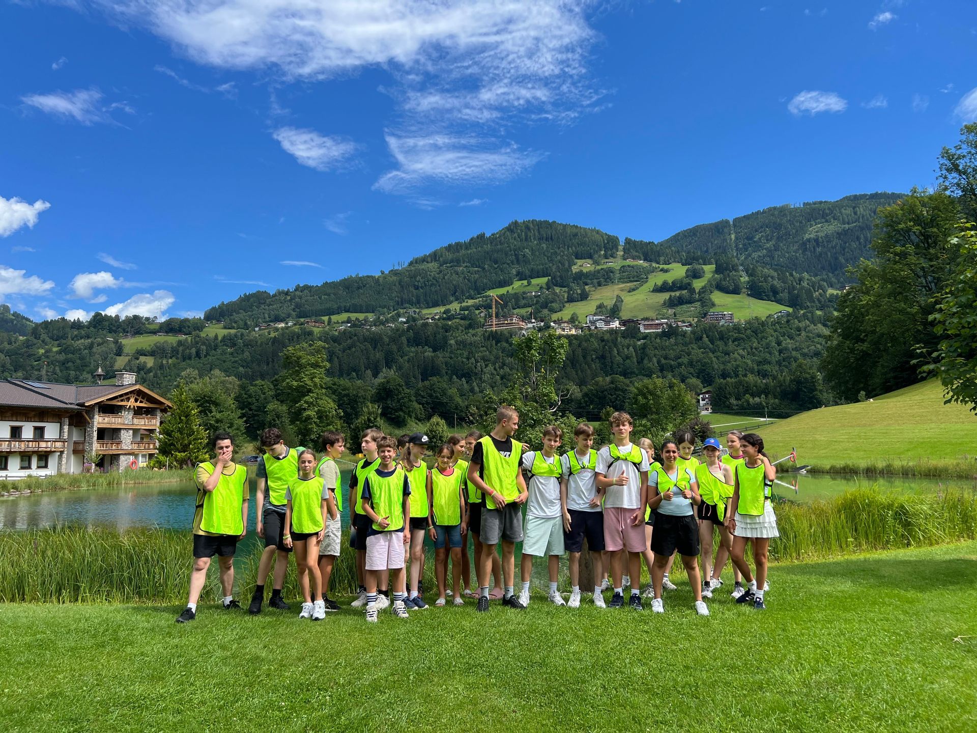 Eine Gruppe von Menschen in neonfarbenen Westen steht auf einer Wiese; im Hintergrund sind Berge und ein See zu sehen.