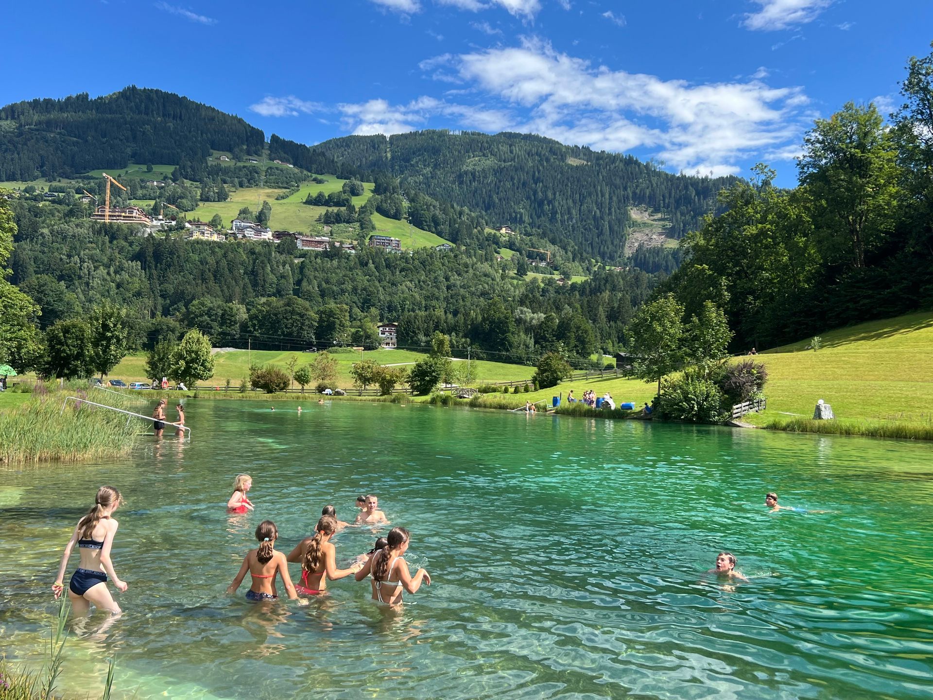 Menschen schwimmen in einem klaren, grünen See vor einer Kulisse aus grünen Hügeln und strahlend blauem Himmel.
