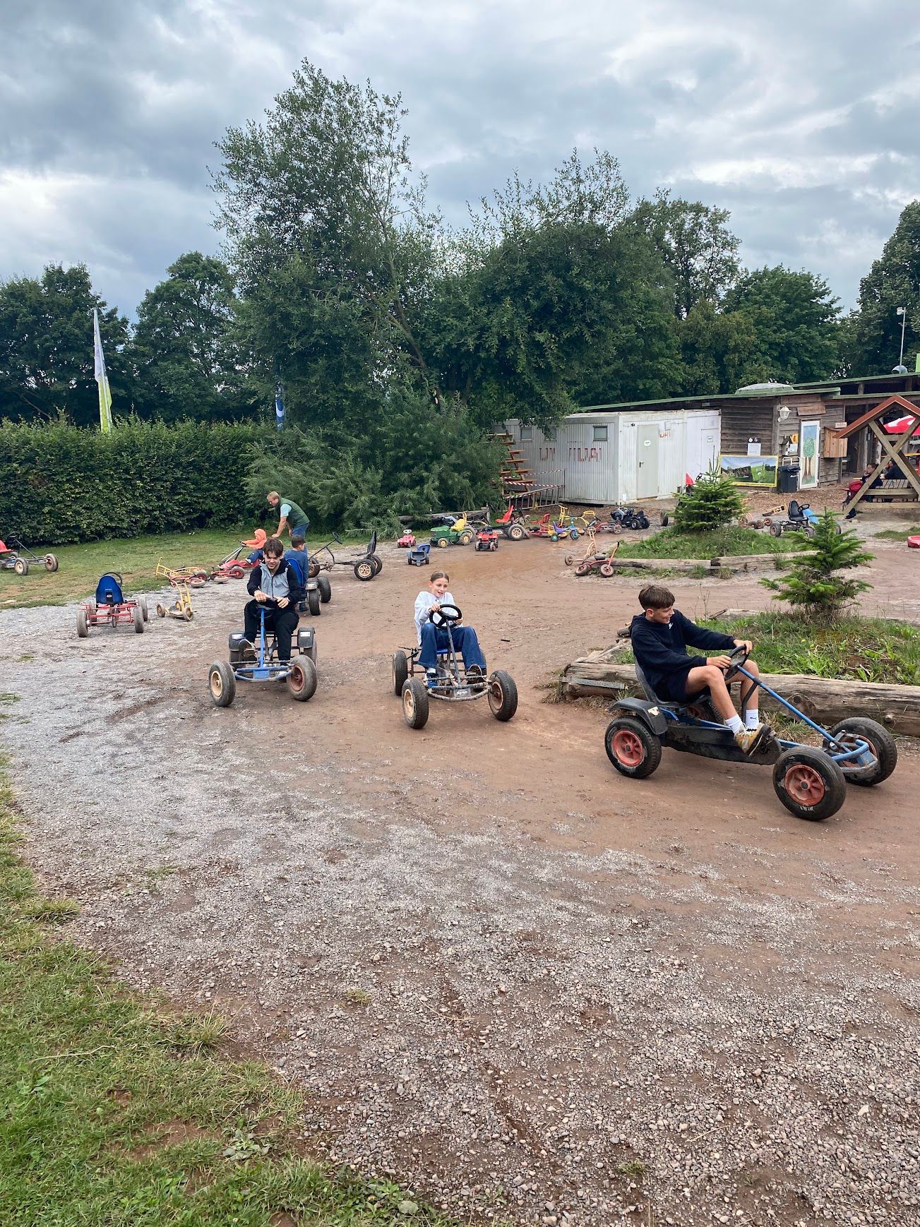 Kinder fahren mit Go-Karts auf einem Schotterweg in der Nähe von Bäumen und einem Gebäude. Bewölkter Himmel.