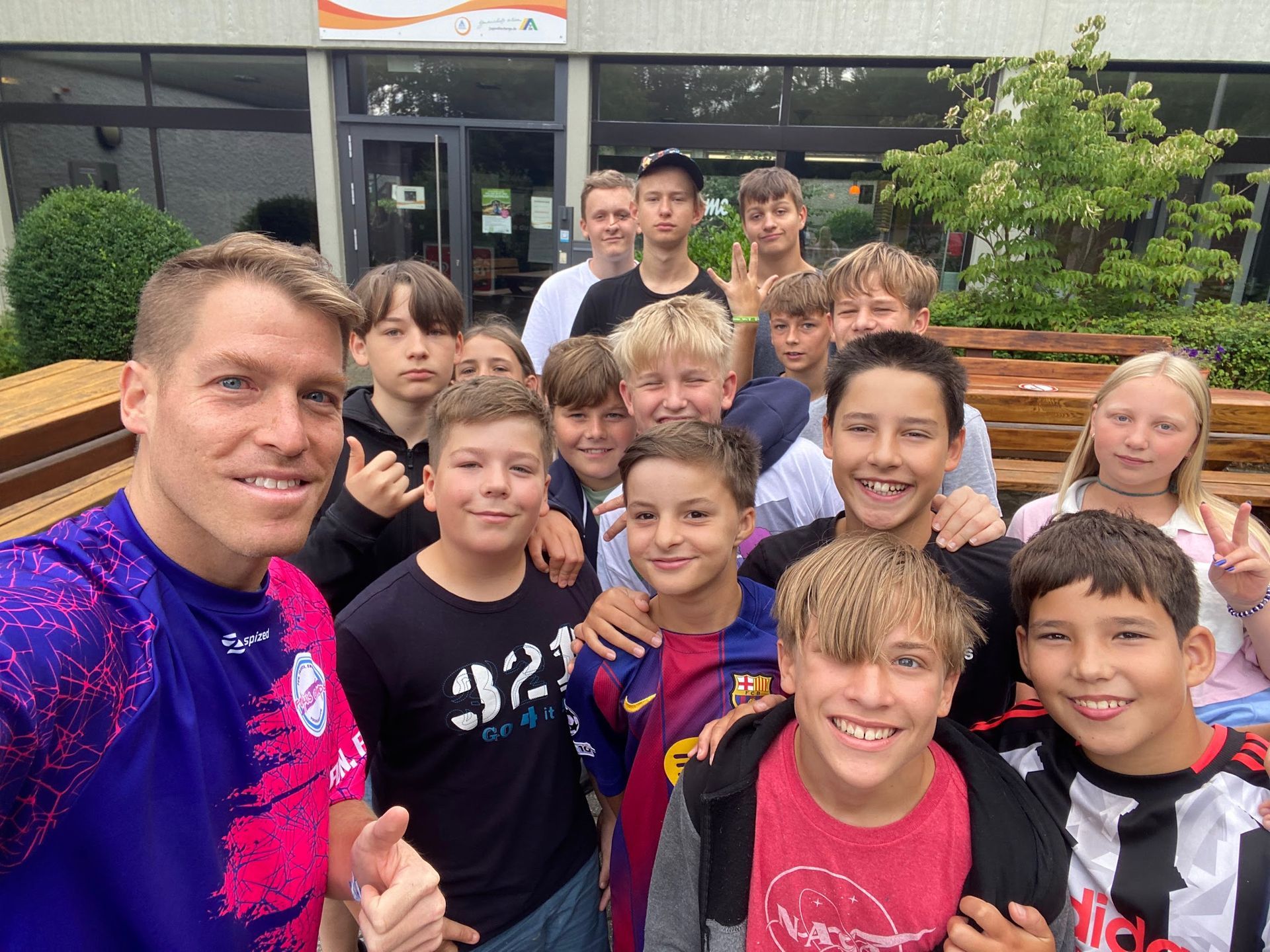 Ein Mann in Sportkleidung macht vor einem Gebäude ein Selfie mit einer Gruppe lächelnder Kinder.