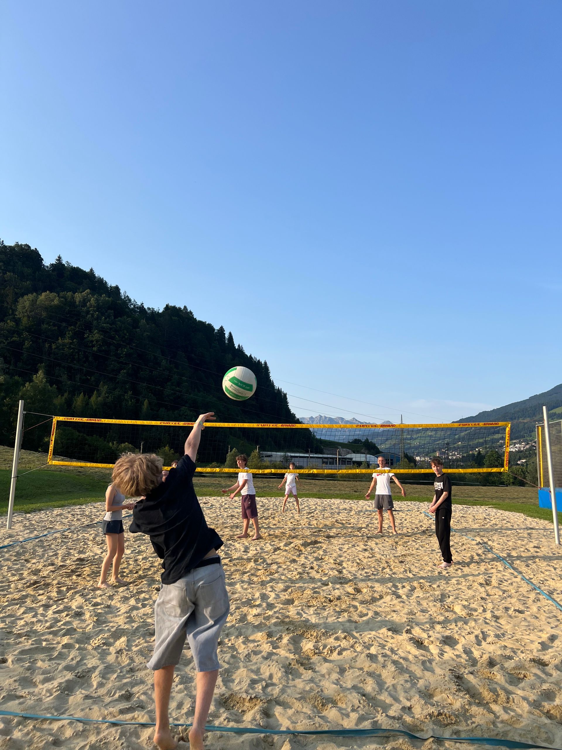 Menschen spielen Volleyball auf einem Sandplatz, der Ball ist in der Luft, im Hintergrund ist ein Netz zu sehen. Berge und blauer Himmel sind erkennbar.