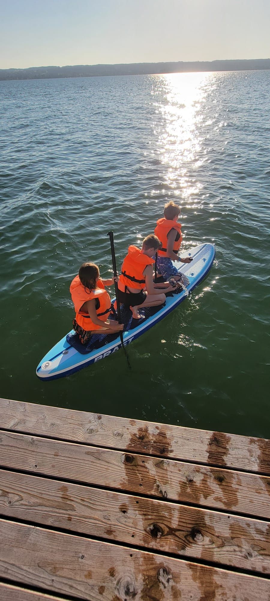 Vier Personen in orangefarbenen Schwimmwesten paddeln auf einem blauen Paddleboard im Wasser. Die Sonne spiegelt sich auf dem Wasser.