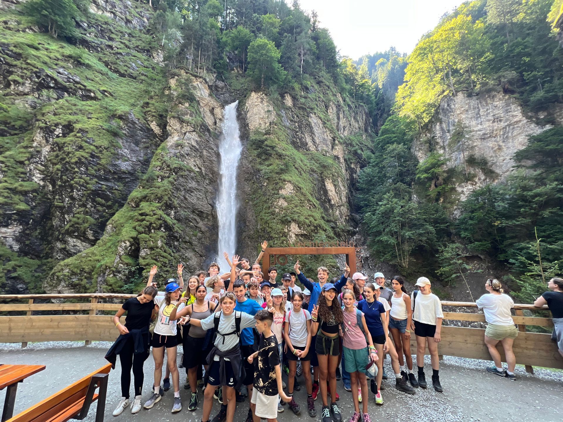 Eine Gruppe von Menschen steht mit erhobenen Armen auf einer Holzplattform vor einem hohen Wasserfall. Grüne Umgebung.