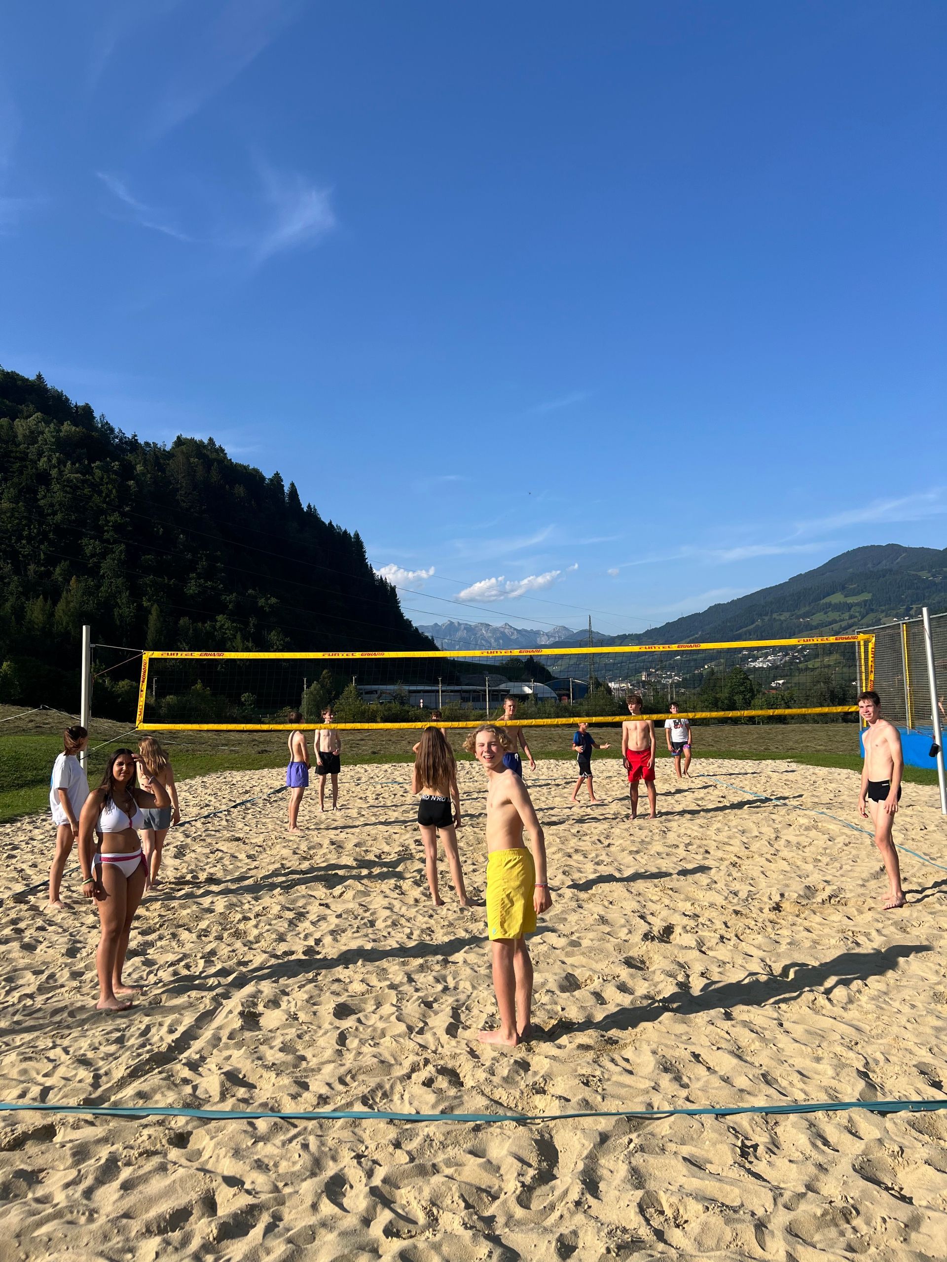 Menschen spielen Volleyball auf einem sandigen Platz, im Hintergrund Berge und blauer Himmel.