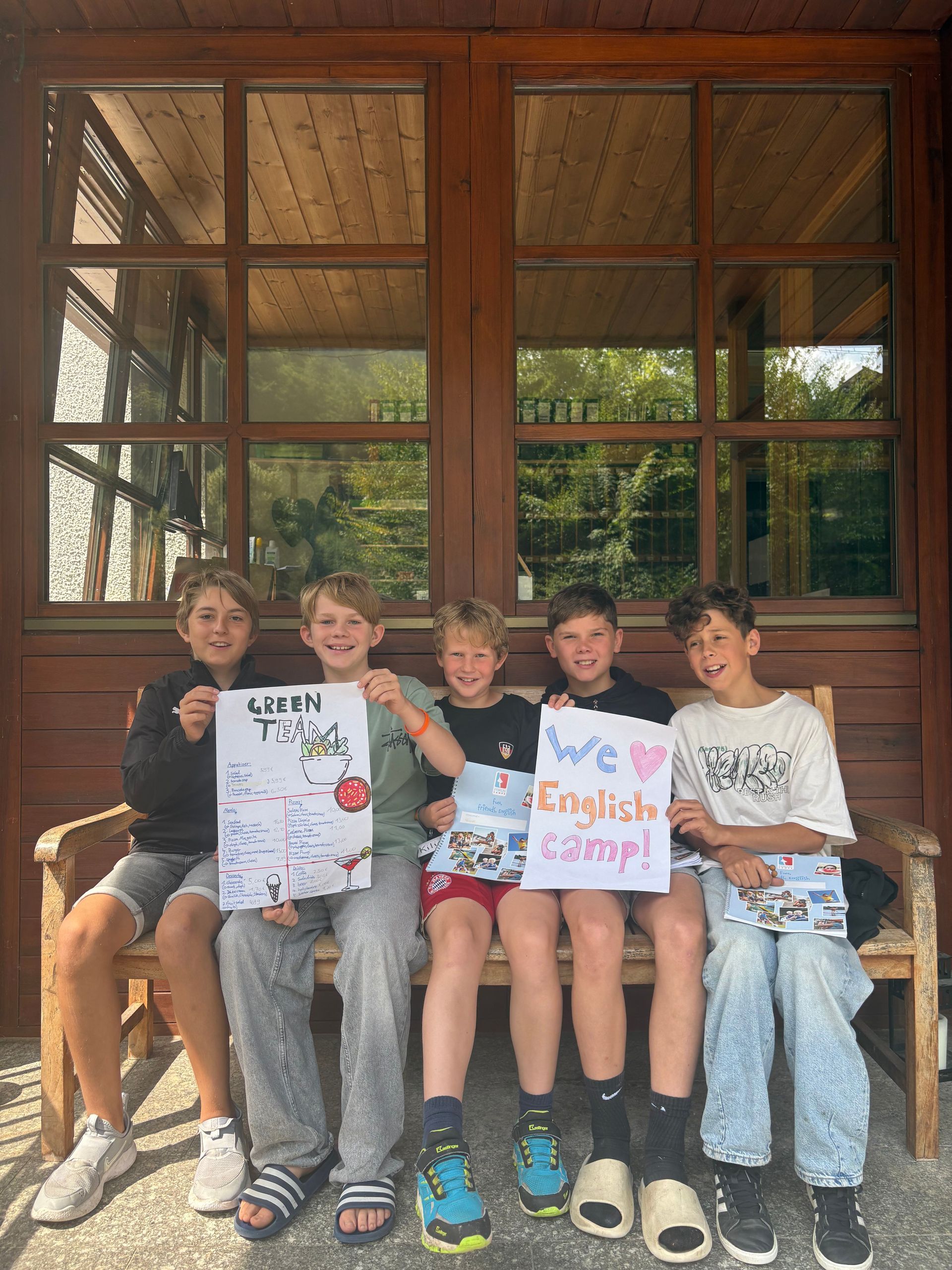 Fünf Jungen sitzen auf einer Holzbank und halten Schilder mit der Aufschrift „Gerry Tiger“ und „Wir lieben das Englischcamp!“ hoch.