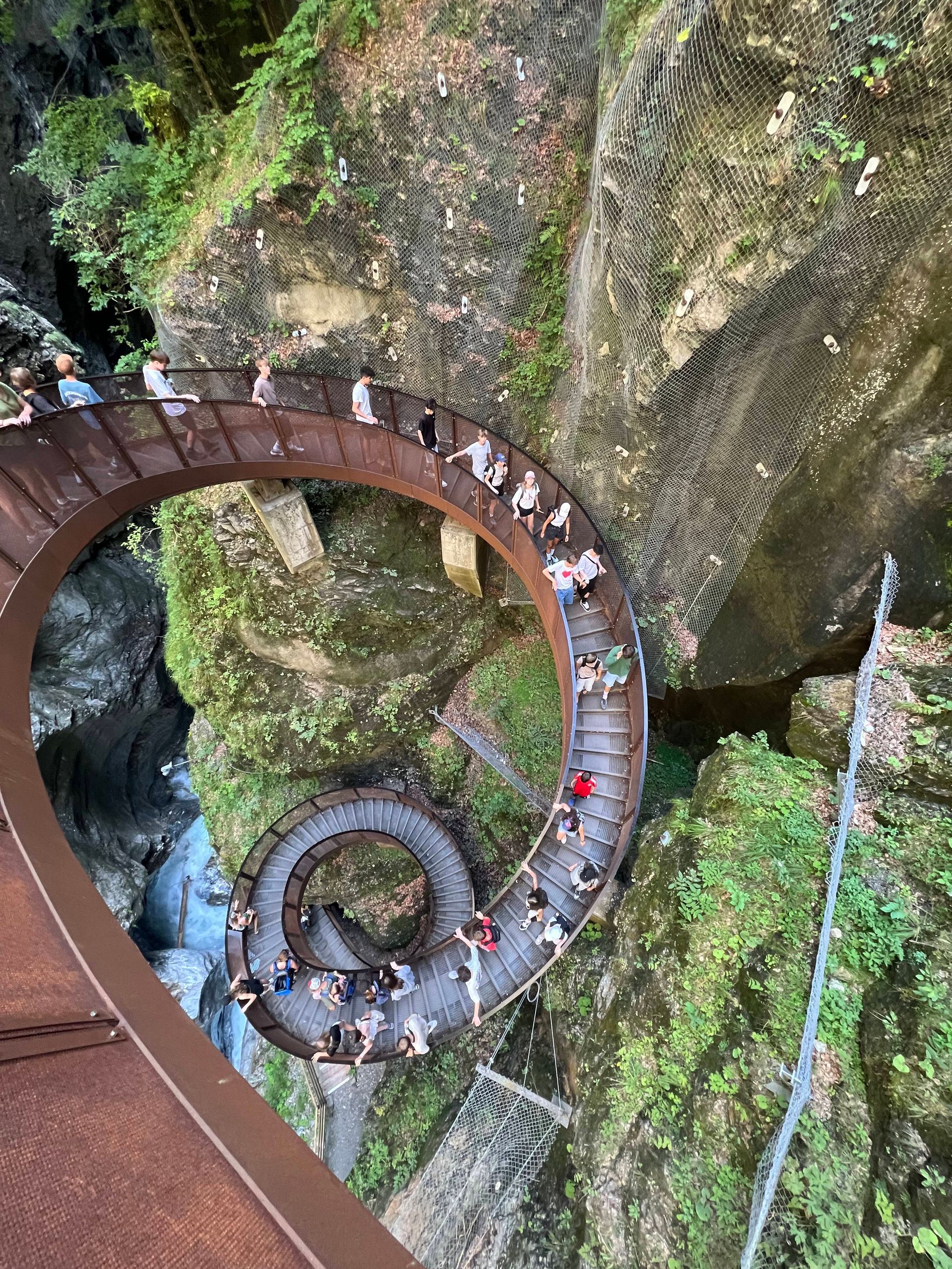 Ein spiralförmiger Steg über einer Schlucht mit vielen Menschen, umgeben von grünem Laubwerk.