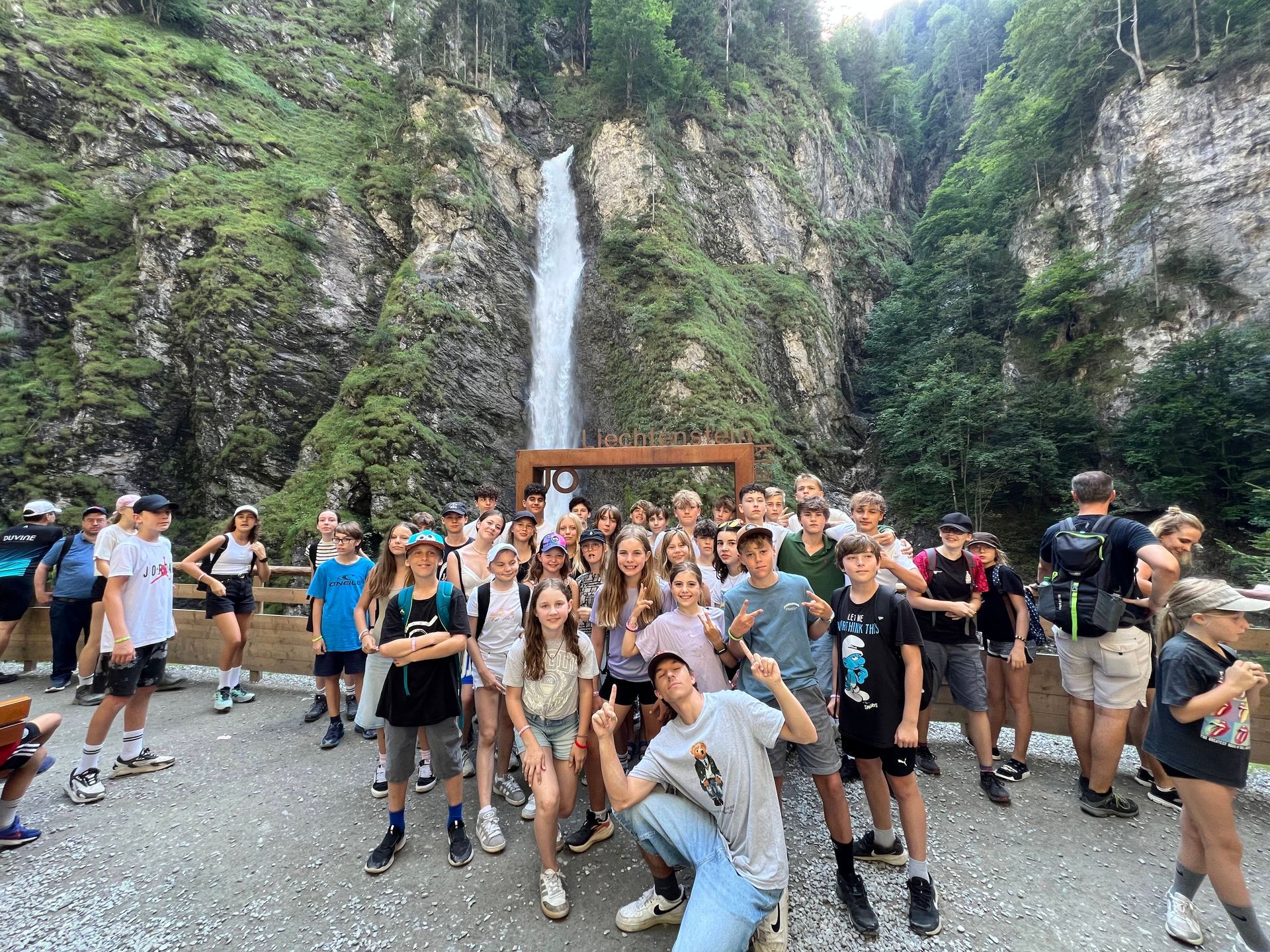 Eine große Gruppe von Menschen posiert vor einem Wasserfall. Sie lächeln und winken.