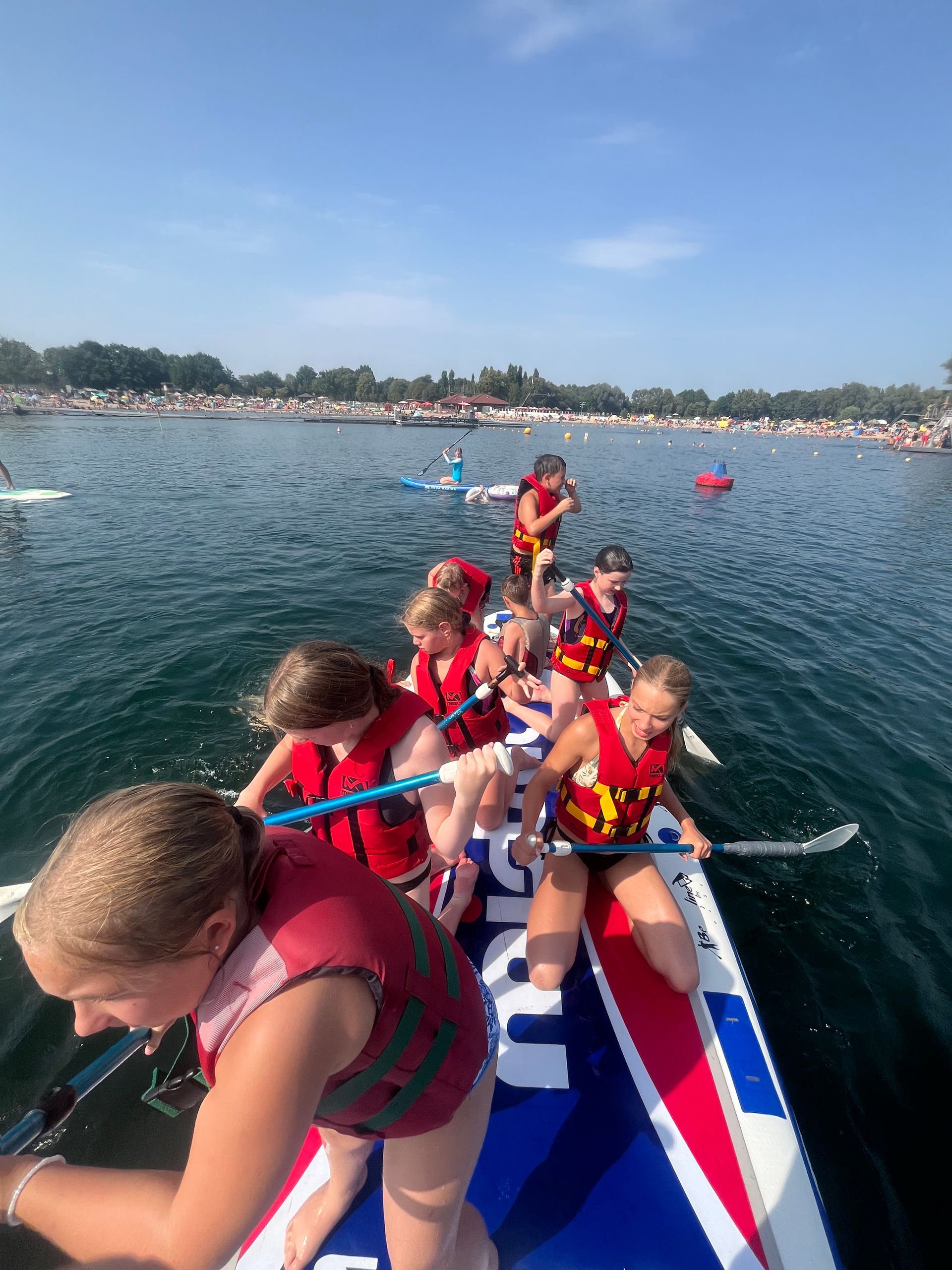 Eine Gruppe Kinder paddelt an einem sonnigen Tag mit Schwimmwesten auf einem großen Paddleboard auf einem See.
