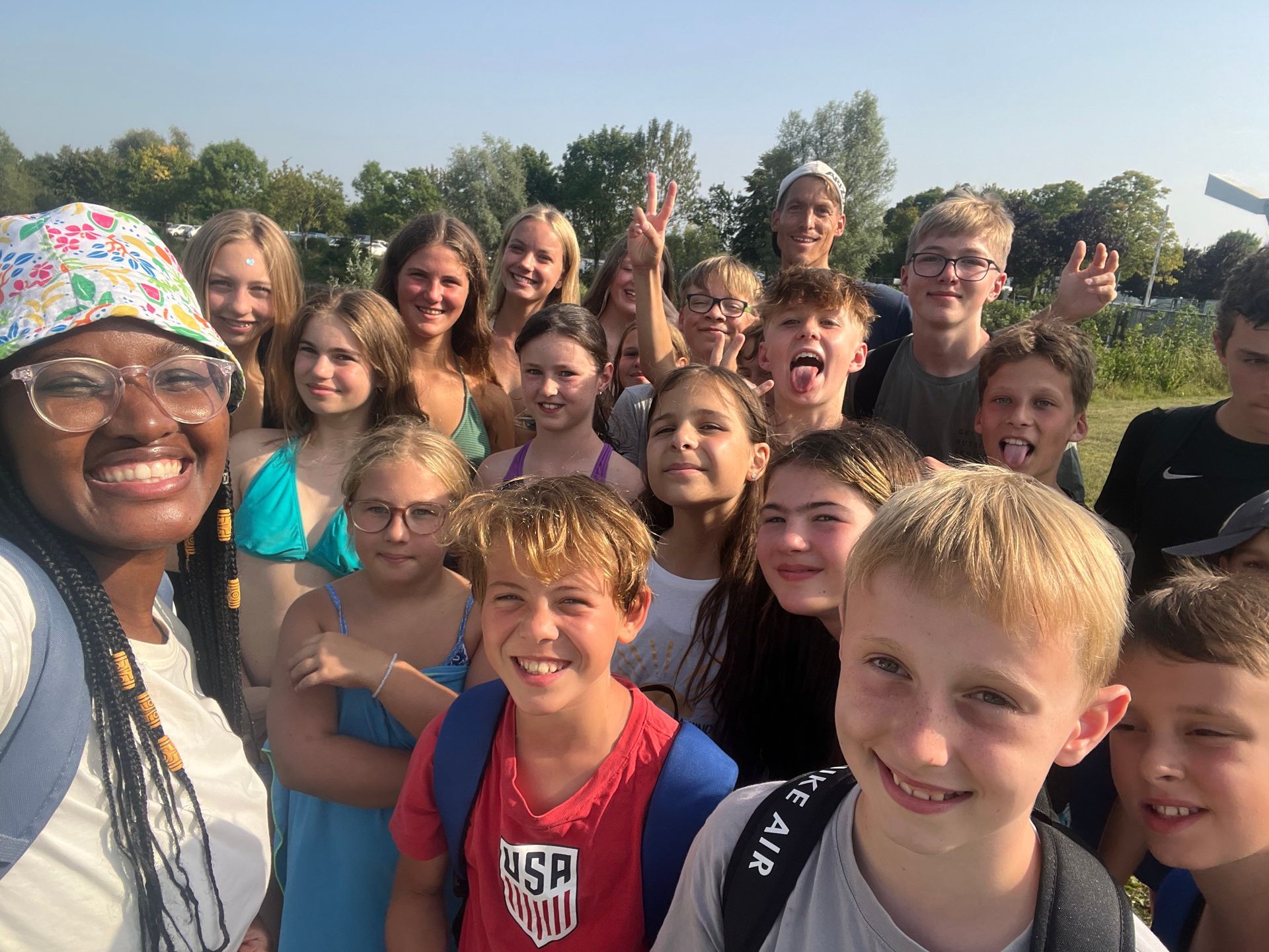 Eine Gruppe lächelnder Kinder und ein Erwachsener posieren im Freien für ein Selfie. Einige tragen Badekleidung.