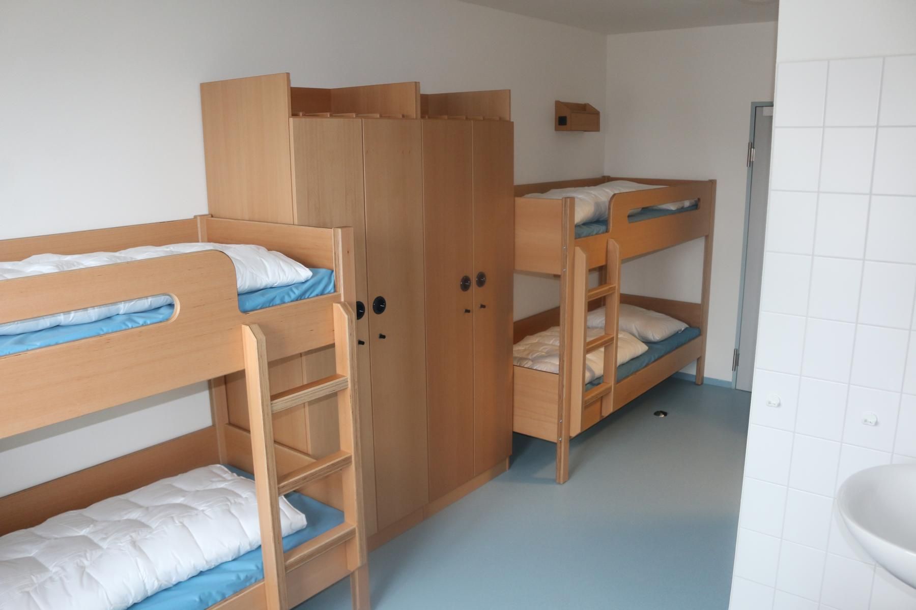 Ein einfaches Studentenwohnheimzimmer mit zwei Etagenbetten und einem großen Holzschrank. Blauer Boden, weiße Wände.