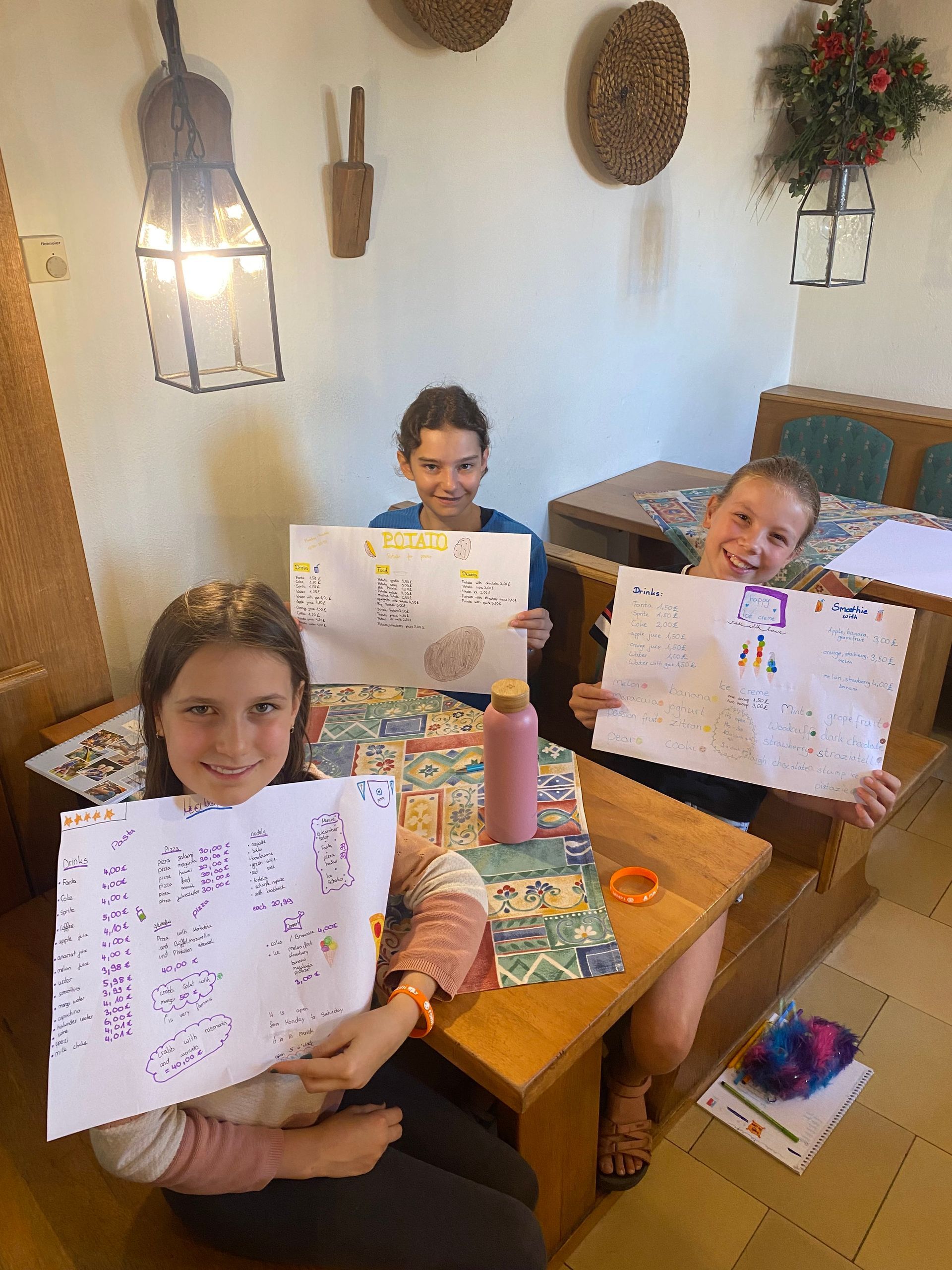 Drei lächelnde Kinder, die Kunstwerke hochhalten, sitzen an einem Holztisch in einem Zimmer mit rustikaler Einrichtung.