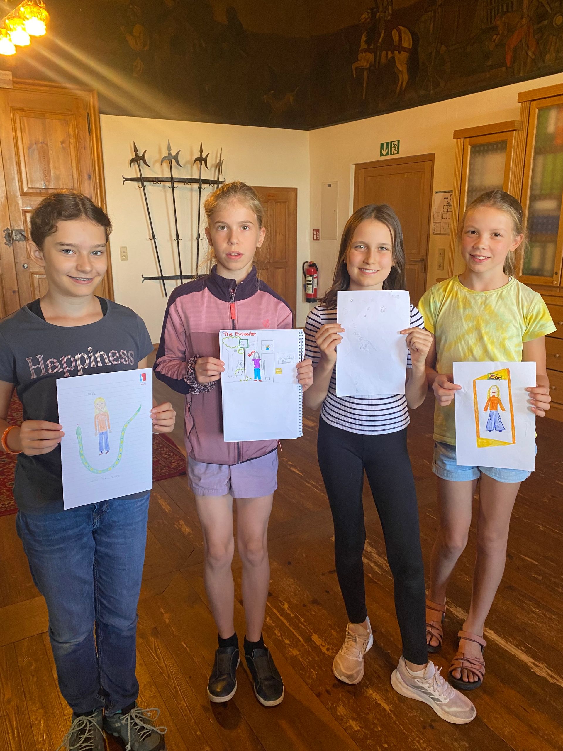 Vier Mädchen halten lächelnd Kunstwerke hoch, drinnen.