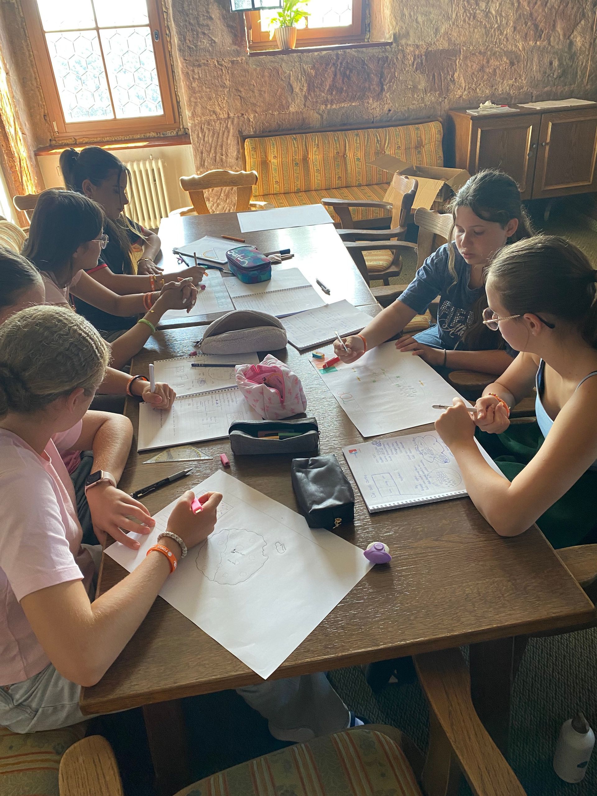 Menschen sitzen an einem Holztisch und zeichnen mit Stiften auf weißes Papier. Innenaufnahme.