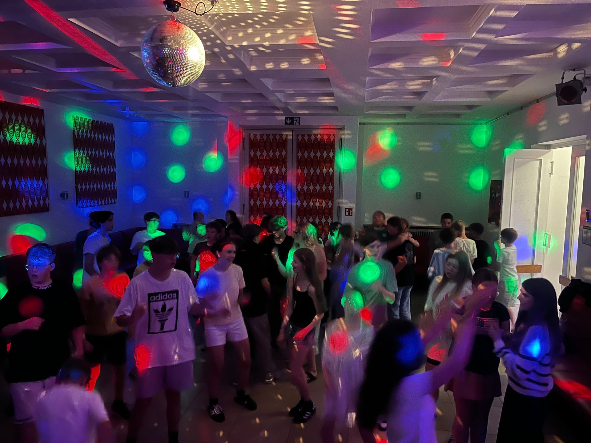 Menschen tanzen auf einer Party in einem Raum mit Discokugel und bunten Lichtern.