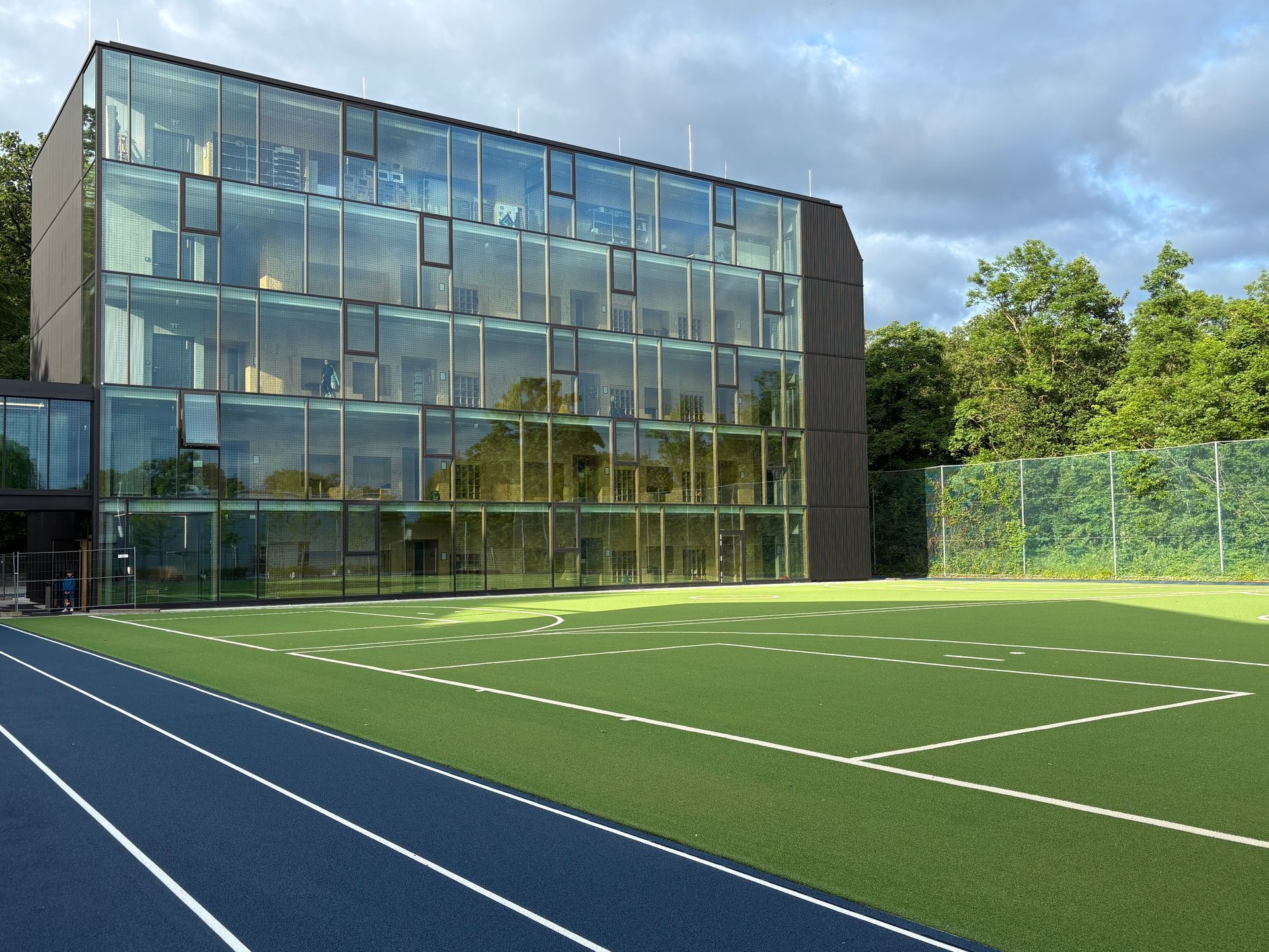 Modernes Glasgebäude mit einem Sportplatz davor und einer blauen Laufbahn an der Seite.