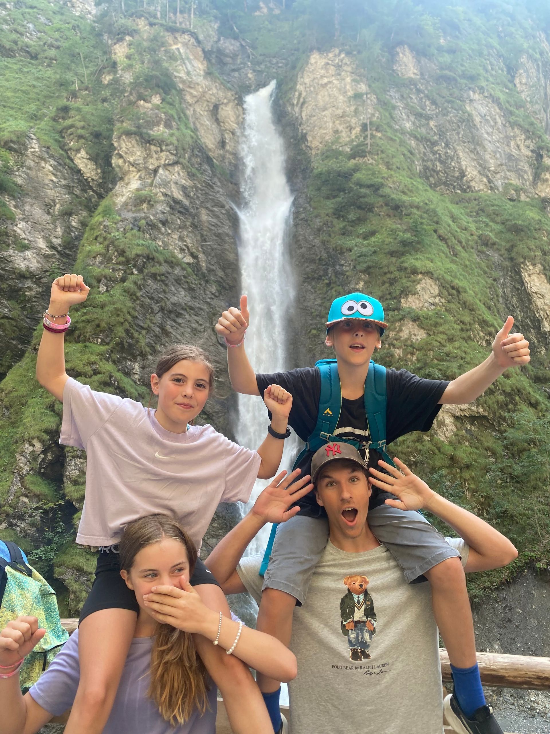 Vier Personen posieren vor einem Wasserfall, zwei sitzen auf den Schultern und jubeln. Üppig grüner Hintergrund.