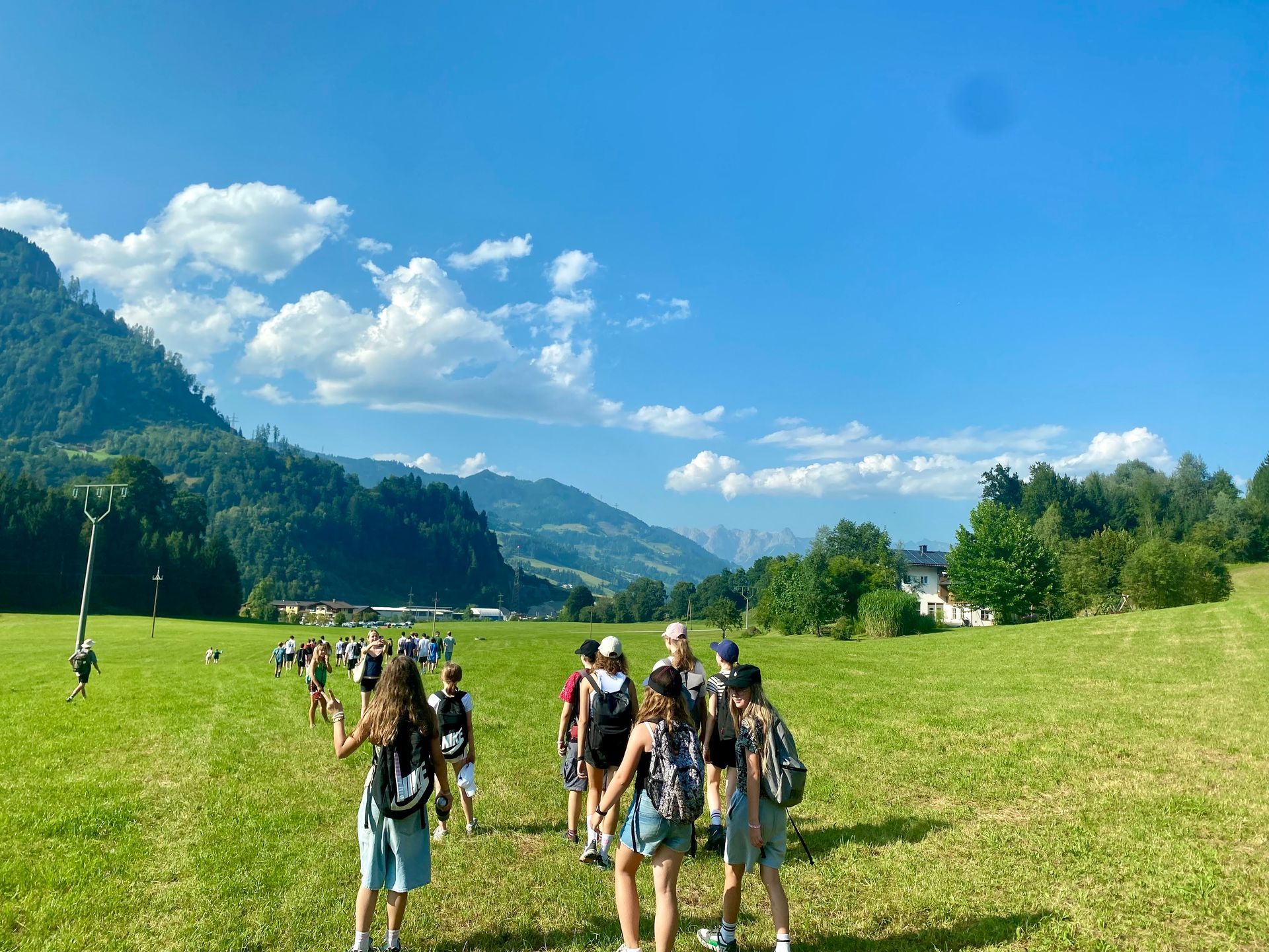 Eine Gruppe von Menschen spaziert unter blauem Himmel durch eine grüne Wiese vor dem Hintergrund von Bergen.