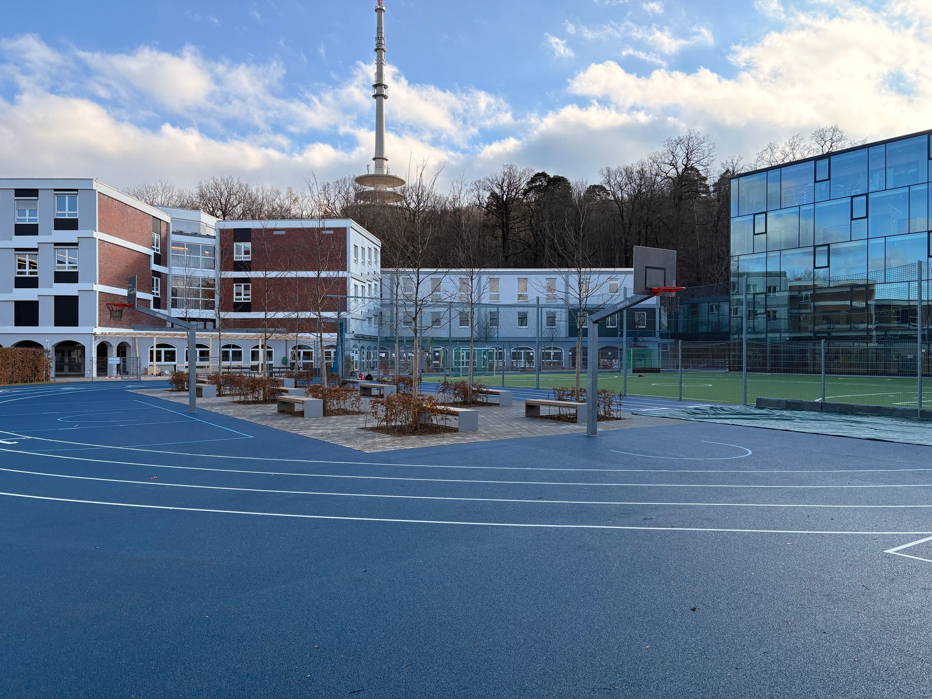 Blauer Schulhof mit Gebäuden, Basketballkorb, Bäumen und einem Turm im Hintergrund.