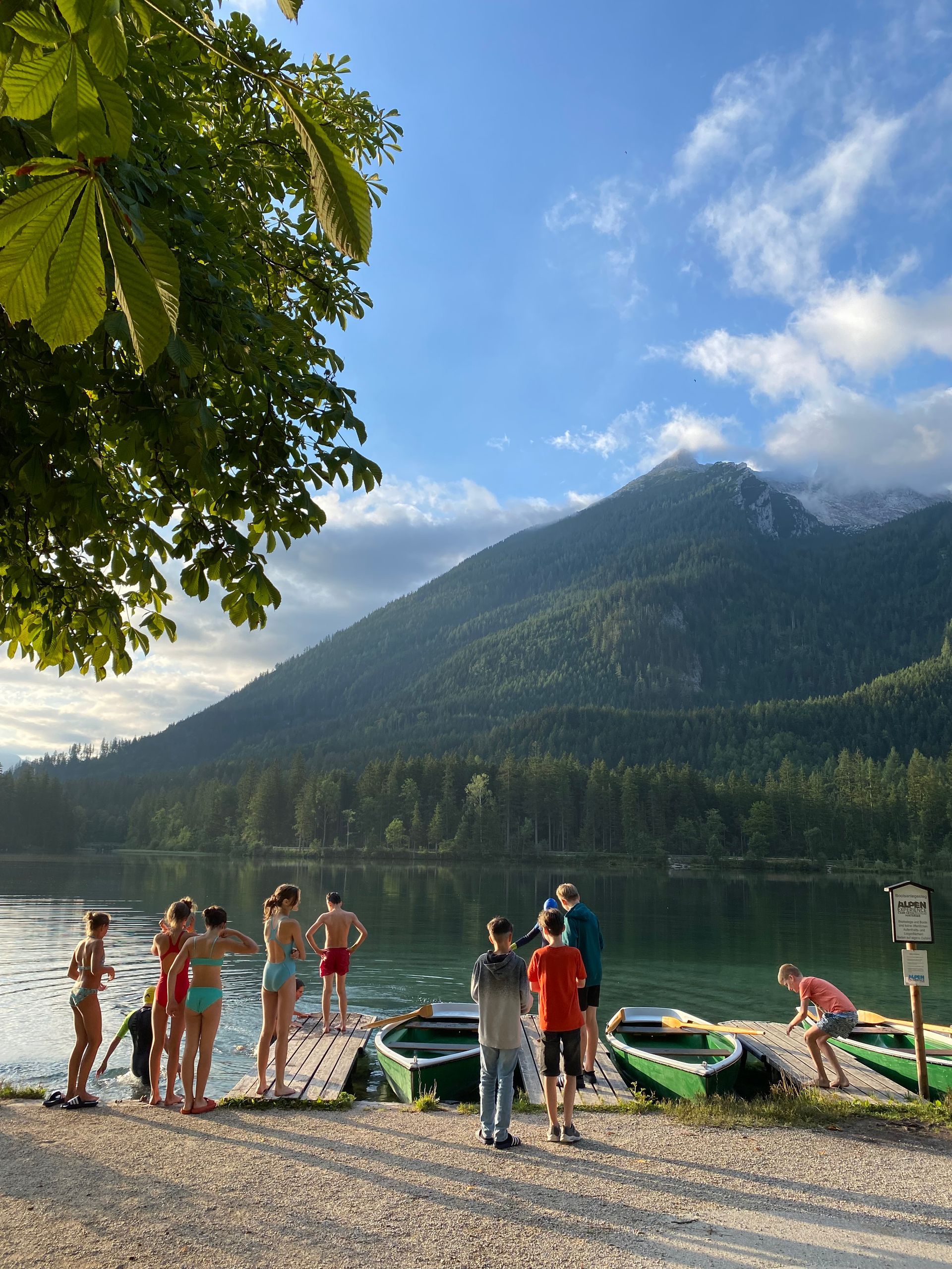 Menschen an einem Seeufer vor einer Bergkulisse. Boote liegen in der Nähe. Einige waten ins Wasser. Blauer Himmel, grüne Bäume.