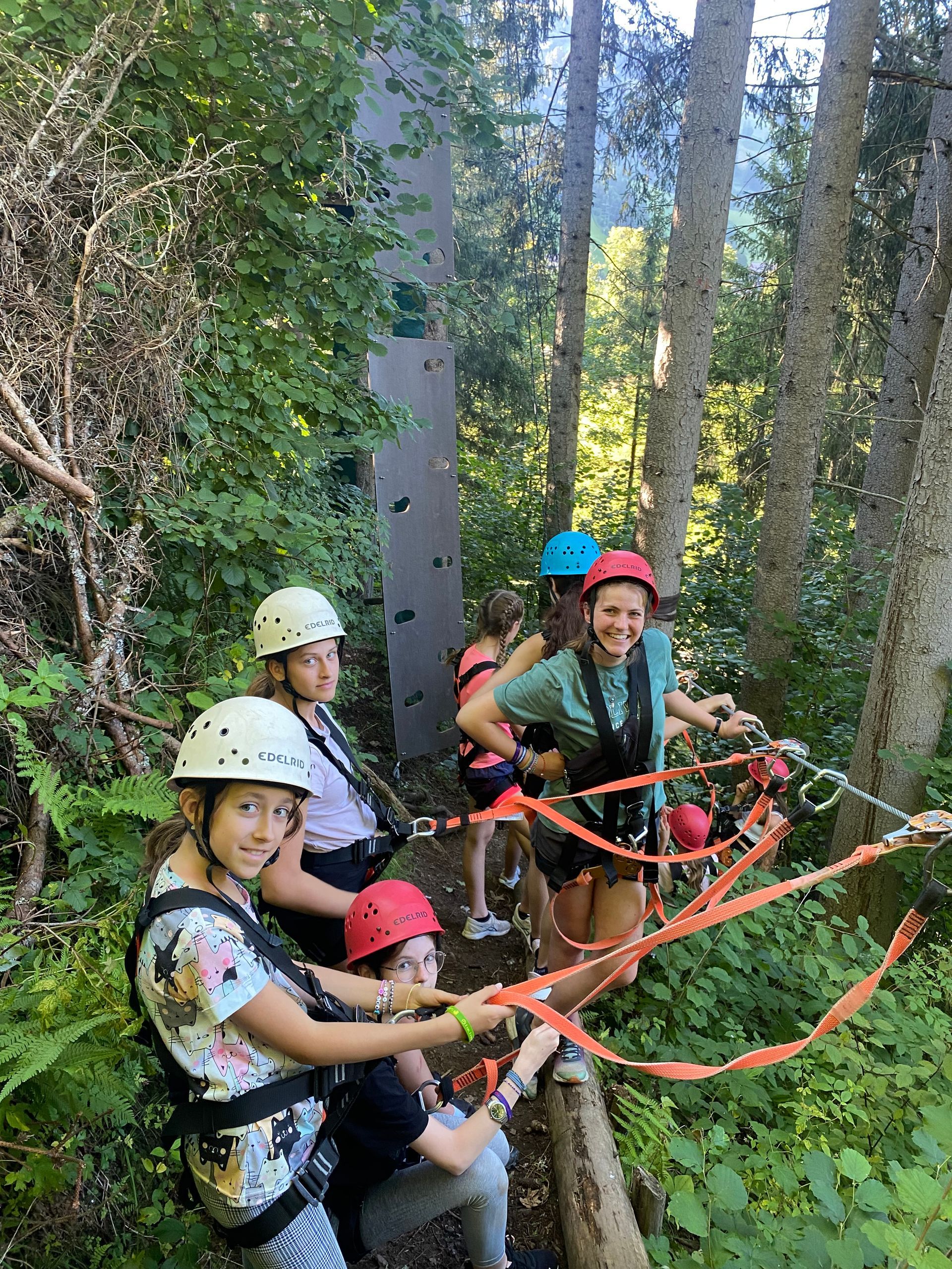 Kinder in Klettergurten auf einem Hochseilgarten; im Hintergrund eine Kletterwand. Waldkulisse, sonniger Tag.