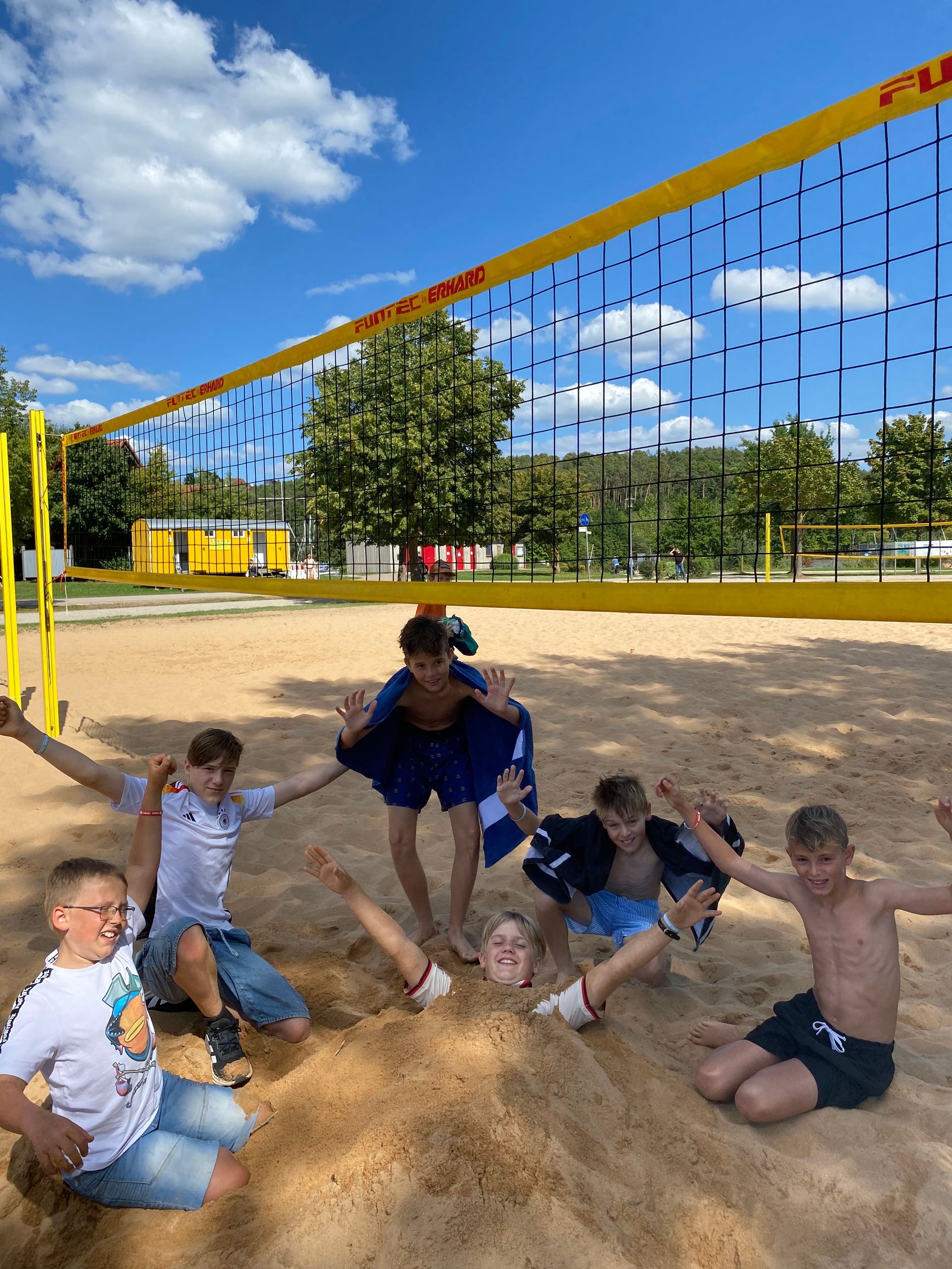 Jungen posieren auf einem sandigen Volleyballfeld unter blauem Himmel. Einige tragen Shorts und T-Shirts.