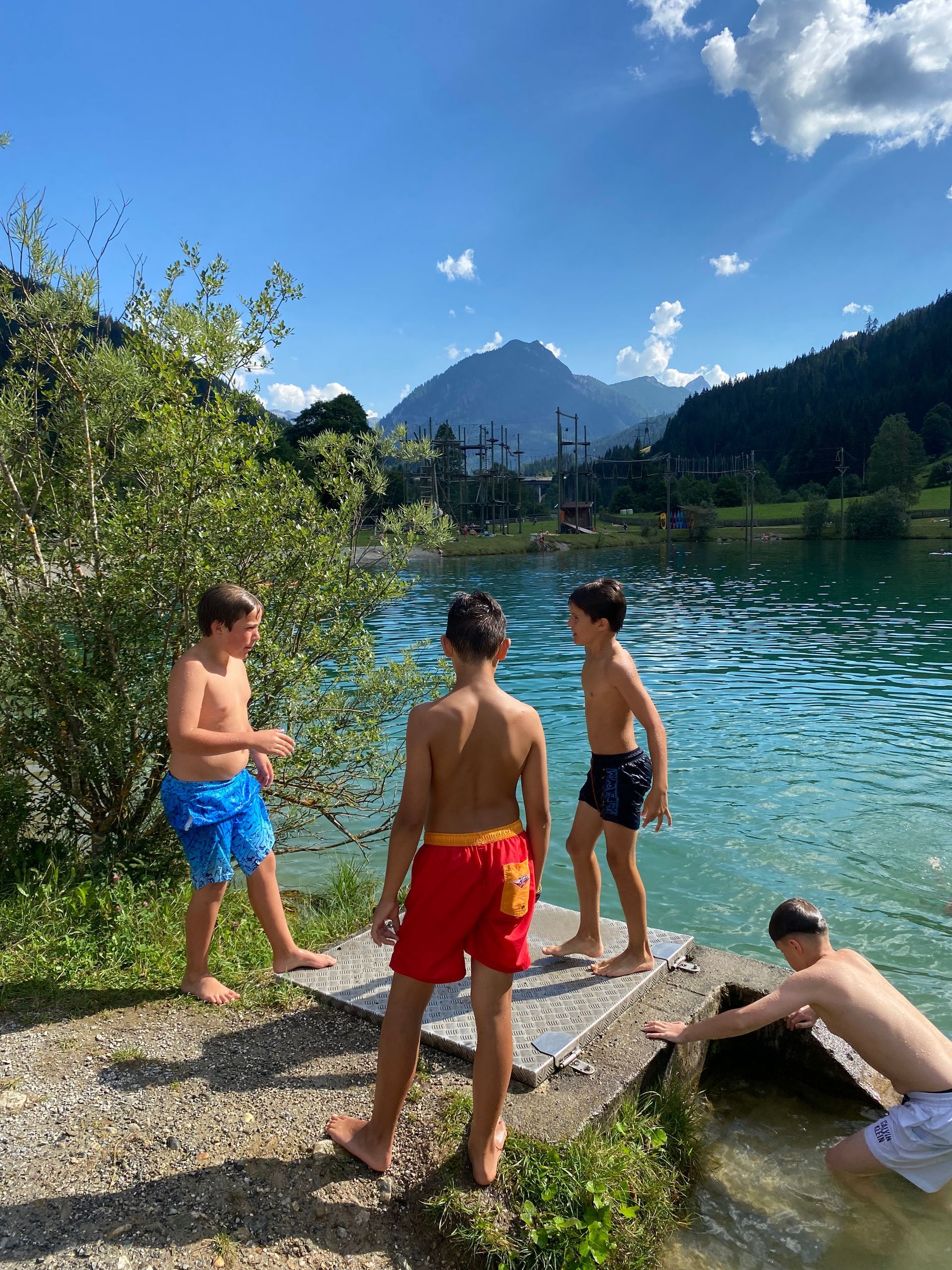 Vier Jungen an einem See, zwei von ihnen in Badehosen. Berge und Bäume im Hintergrund, sonniger Tag.